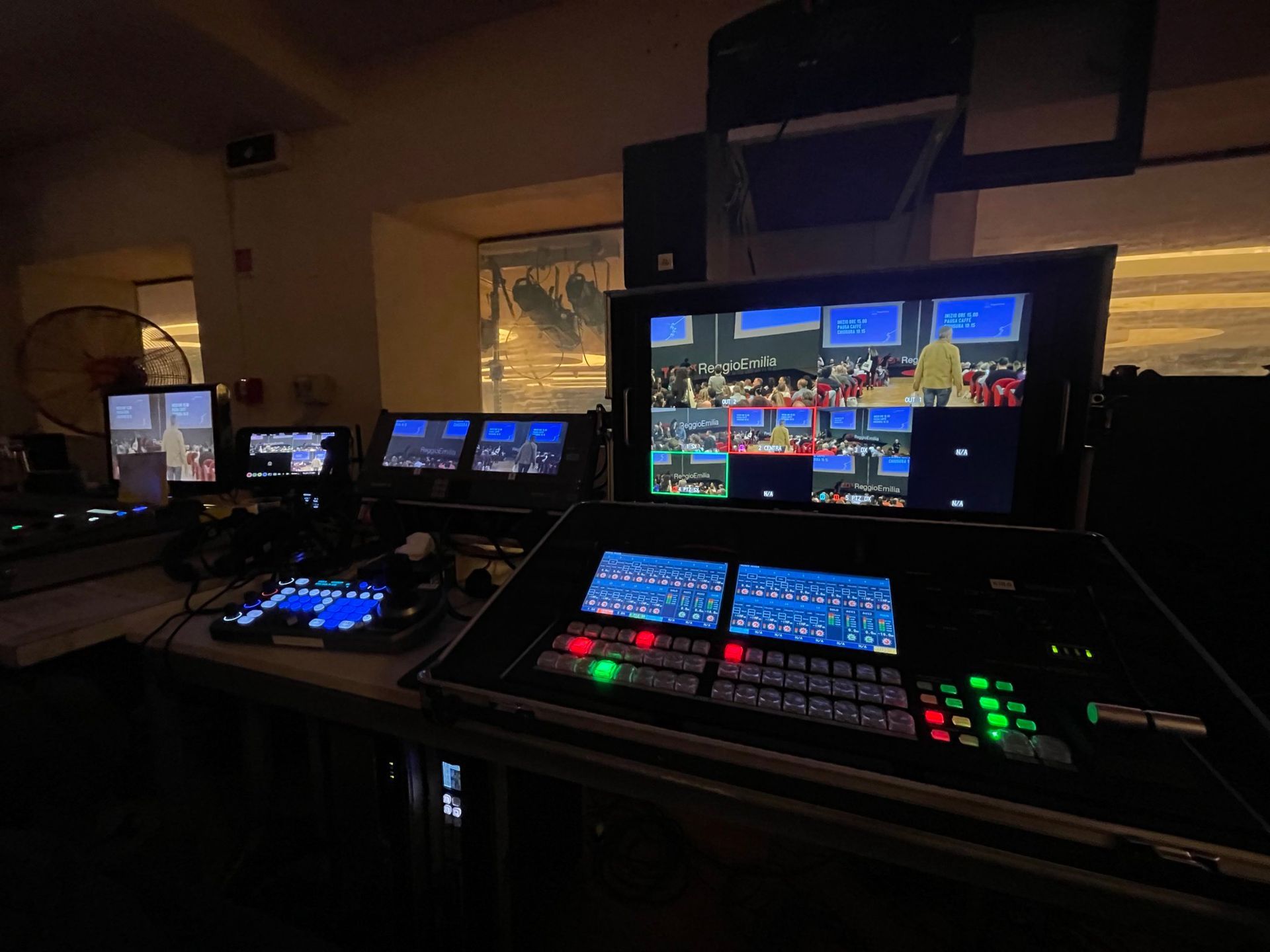 Sala di controllo con più schermi che mostrano un evento in diretta. L'attrezzatura include switcher e monitor. Interno in penombra con vista dalle finestre.