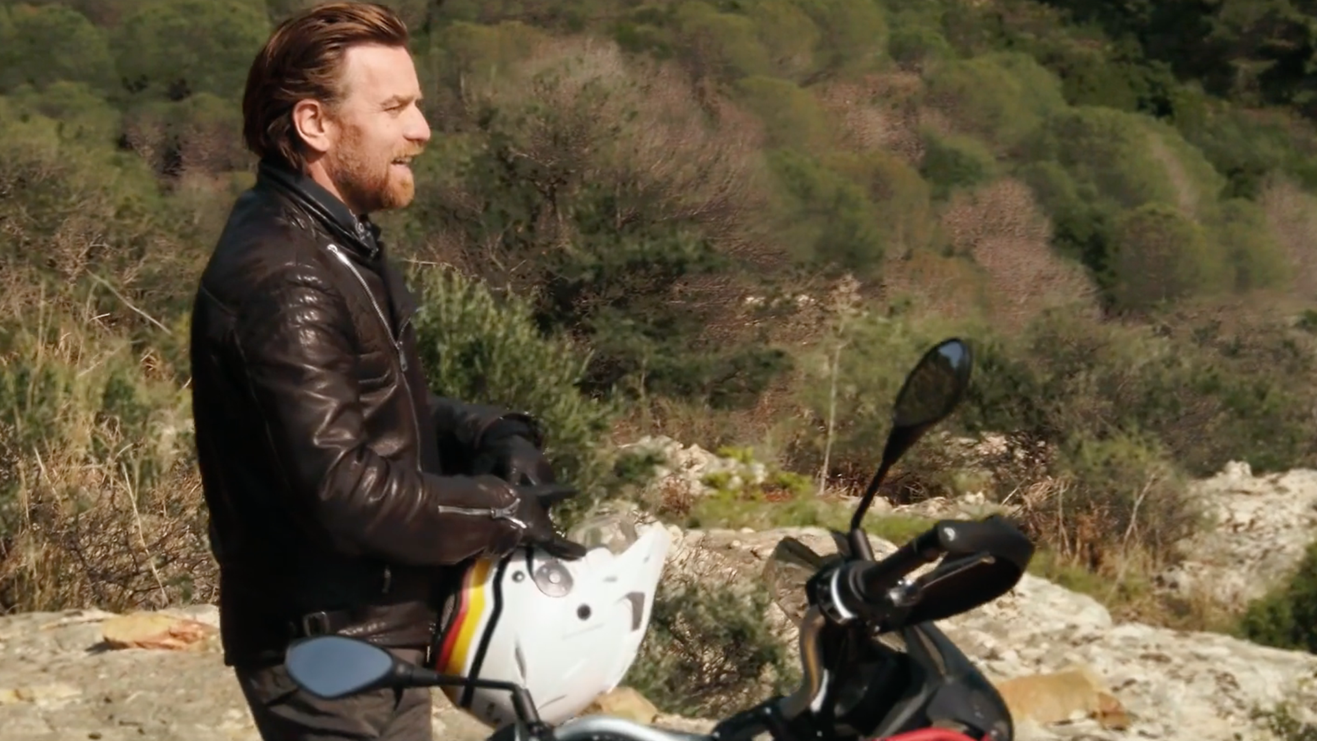 Man in leather jacket by a motorcycle, holding a helmet, overlooking a rocky landscape.