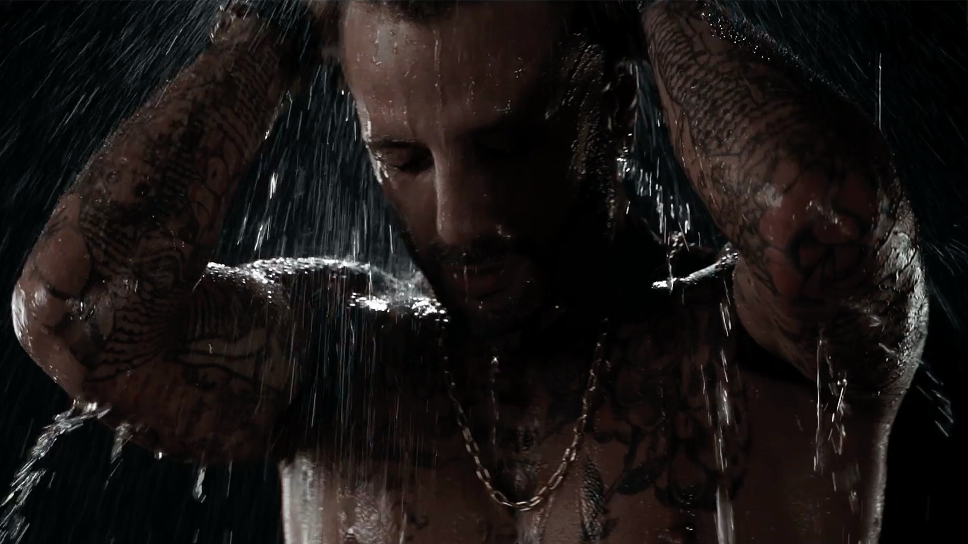 Man in shower, water cascading. He has tattoos and a gold chain, dark background.