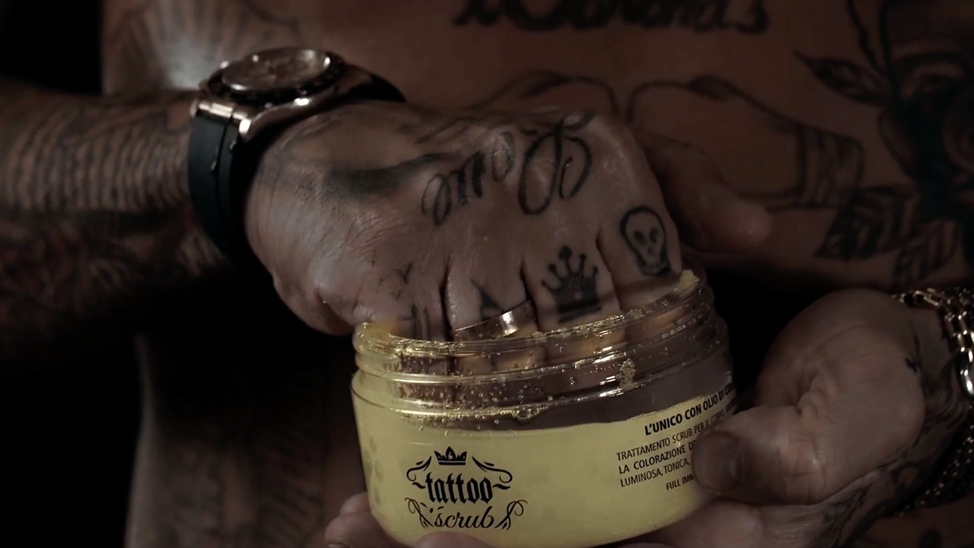 Tattooed hand holding a jar of tattoo balm. Gold watch and jewelry visible.