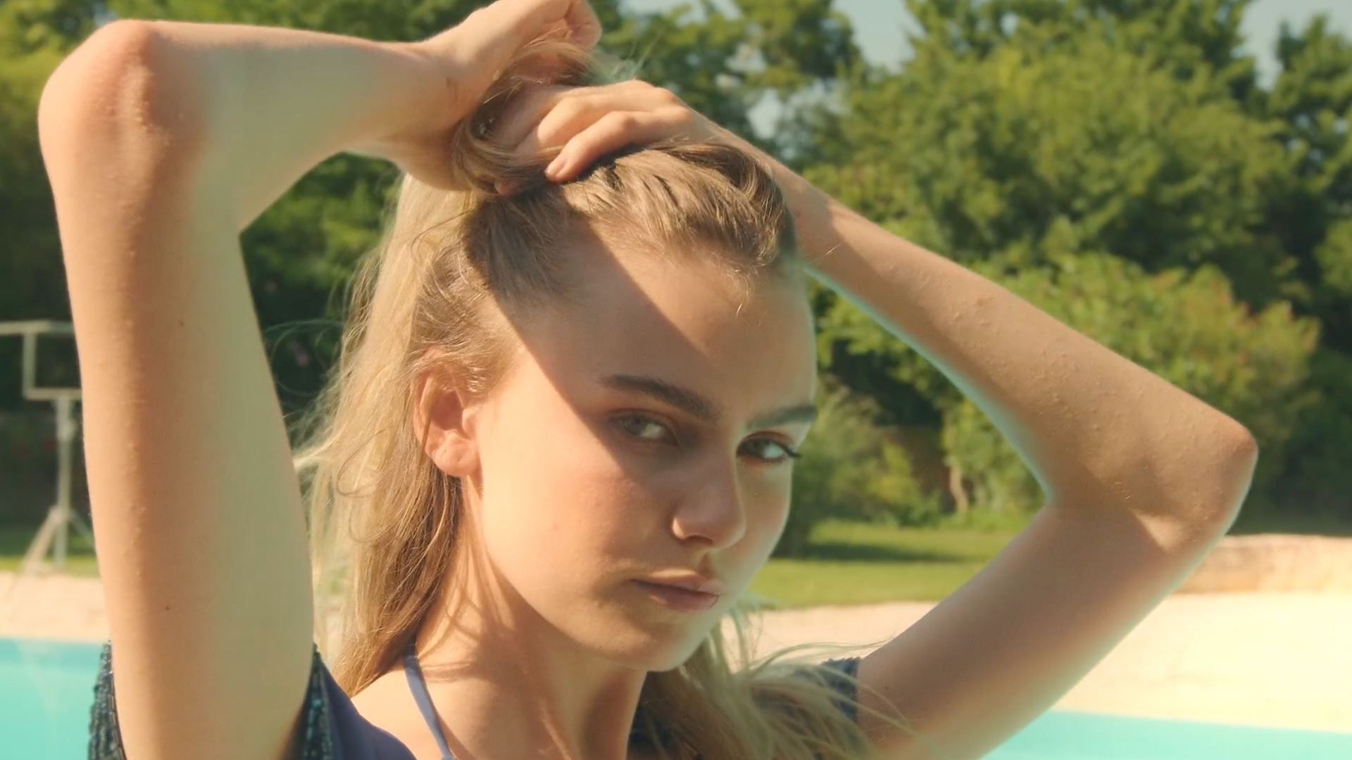 Donna con i capelli biondi, che li tiene raccolti, guarda verso la telecamera. Piscina sullo sfondo.
