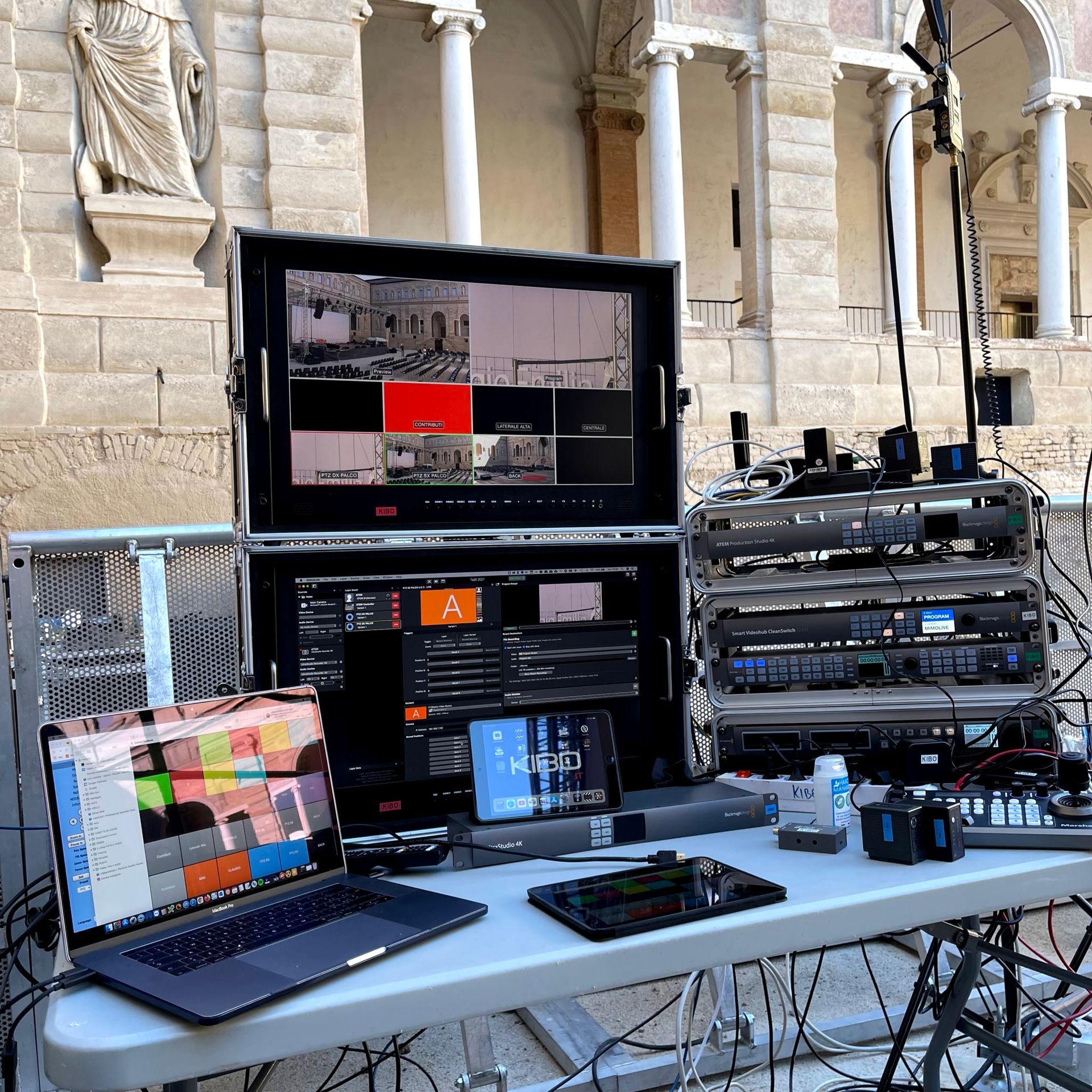 Attrezzatura per trasmissioni all'aperto: monitor, mixer audio, laptop, tablet su un tavolo, con un edificio sullo sfondo.