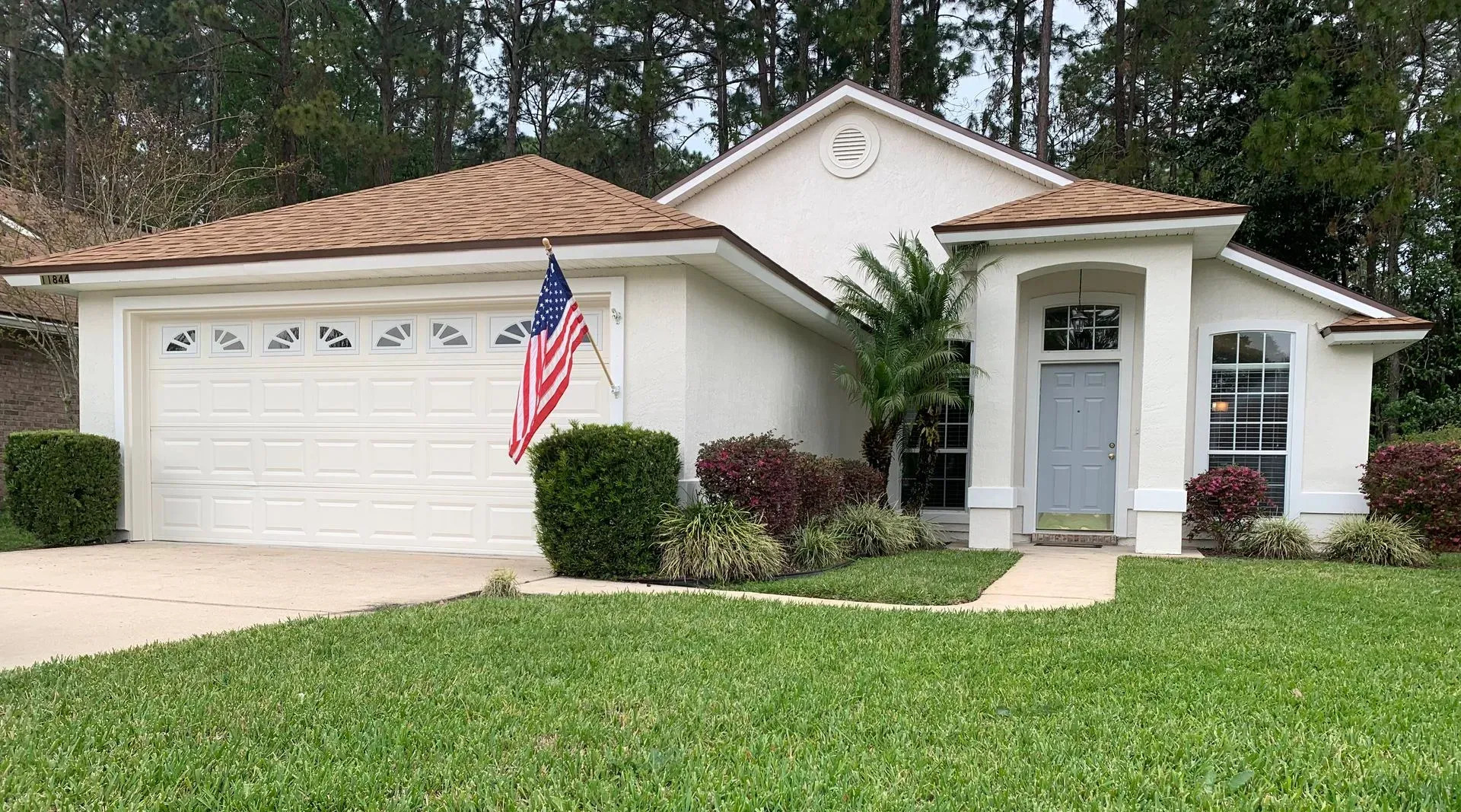 Single-story cream house with tan roof, white garage, front porch, and American flag in a green lawn.
