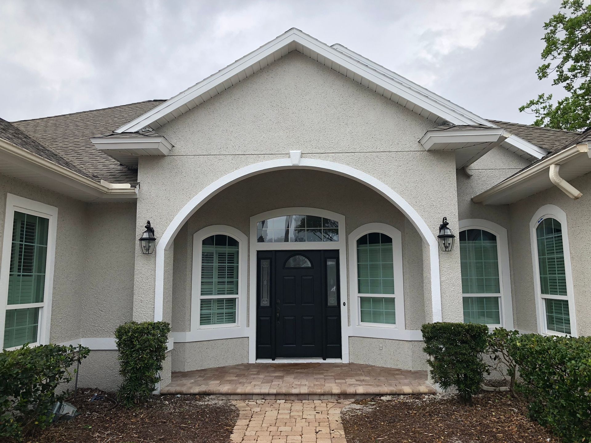 Front entrance of a light gray stucco house with arched windows, black door, and brick steps