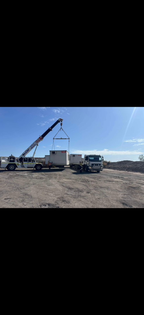 A crane lifting a box, likely for transport, in a dusty outdoor setting under a blue sky.— Fitzroy Towing & Transport in Parkhurst, QLD
