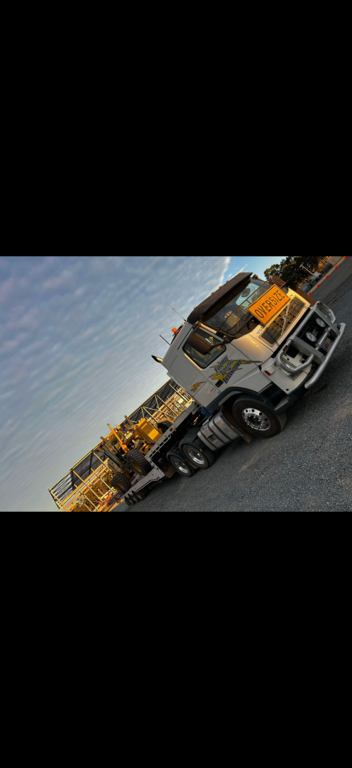 A large white truck carrying construction equipment on a gray road with a cloudy sky.