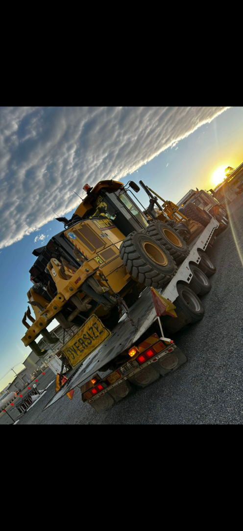 A large yellow construction vehicle is being transported on a trailer on a road at sunset.