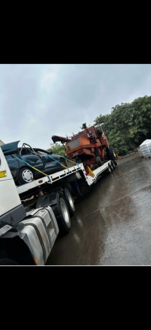 Tow truck carrying a car and what appears to be a large piece of equipment on a wet road. Cloudy sky overhead.