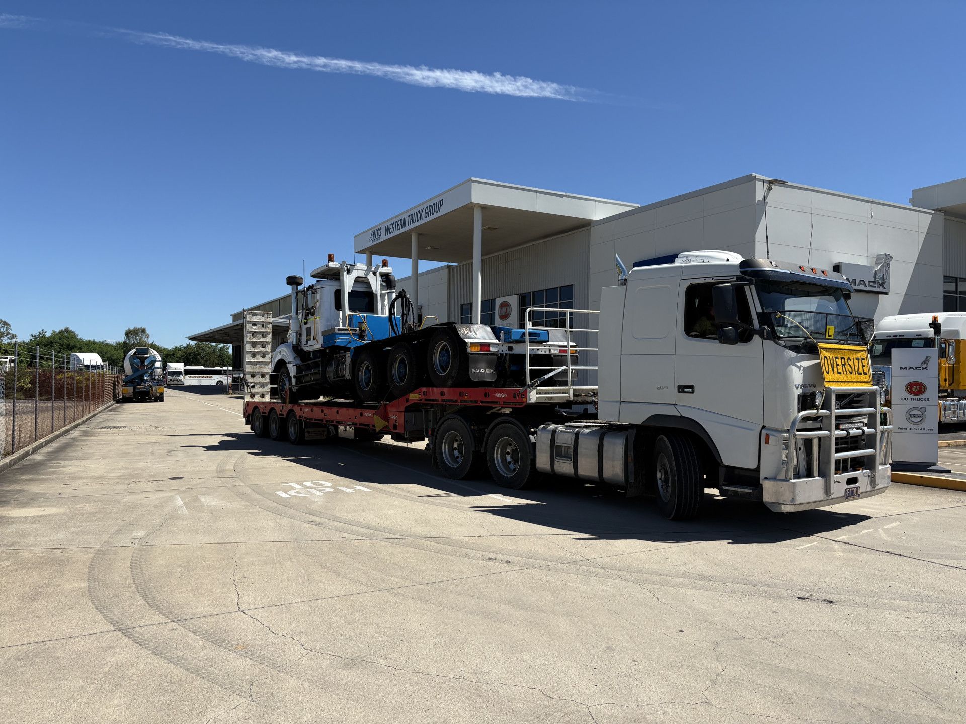 A white truck hauling a blue and white tractor on a flatbed trailer in front of a building on a sunny day.— Fitzroy Towing & Transport in Parkhurst, QLD
