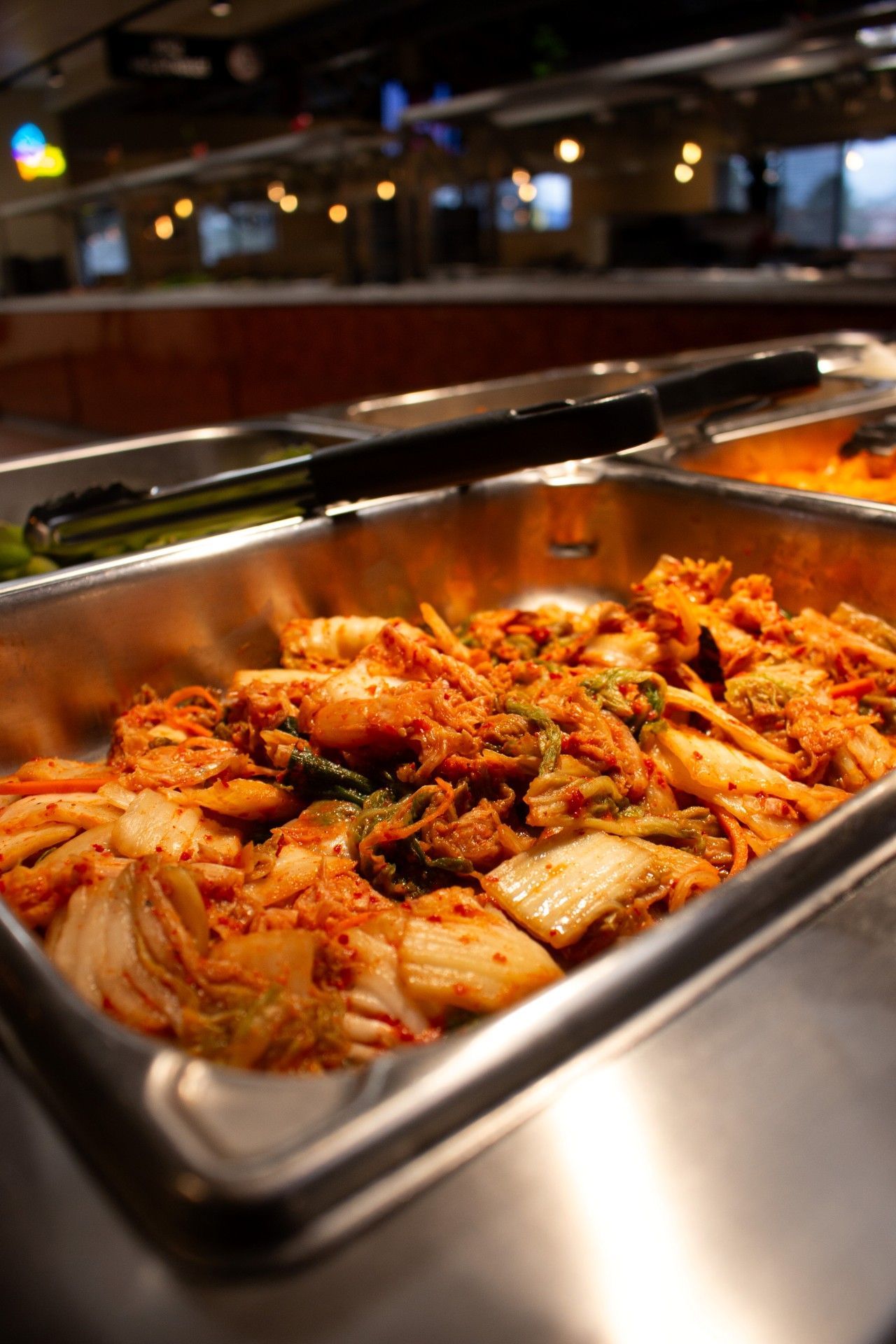 Tray of spicy kimchi in a buffet serving station, with stainless steel pans and warm lighting.