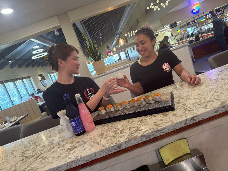 Two people chatting across a restaurant counter, with sushi trays and drinks on the table.