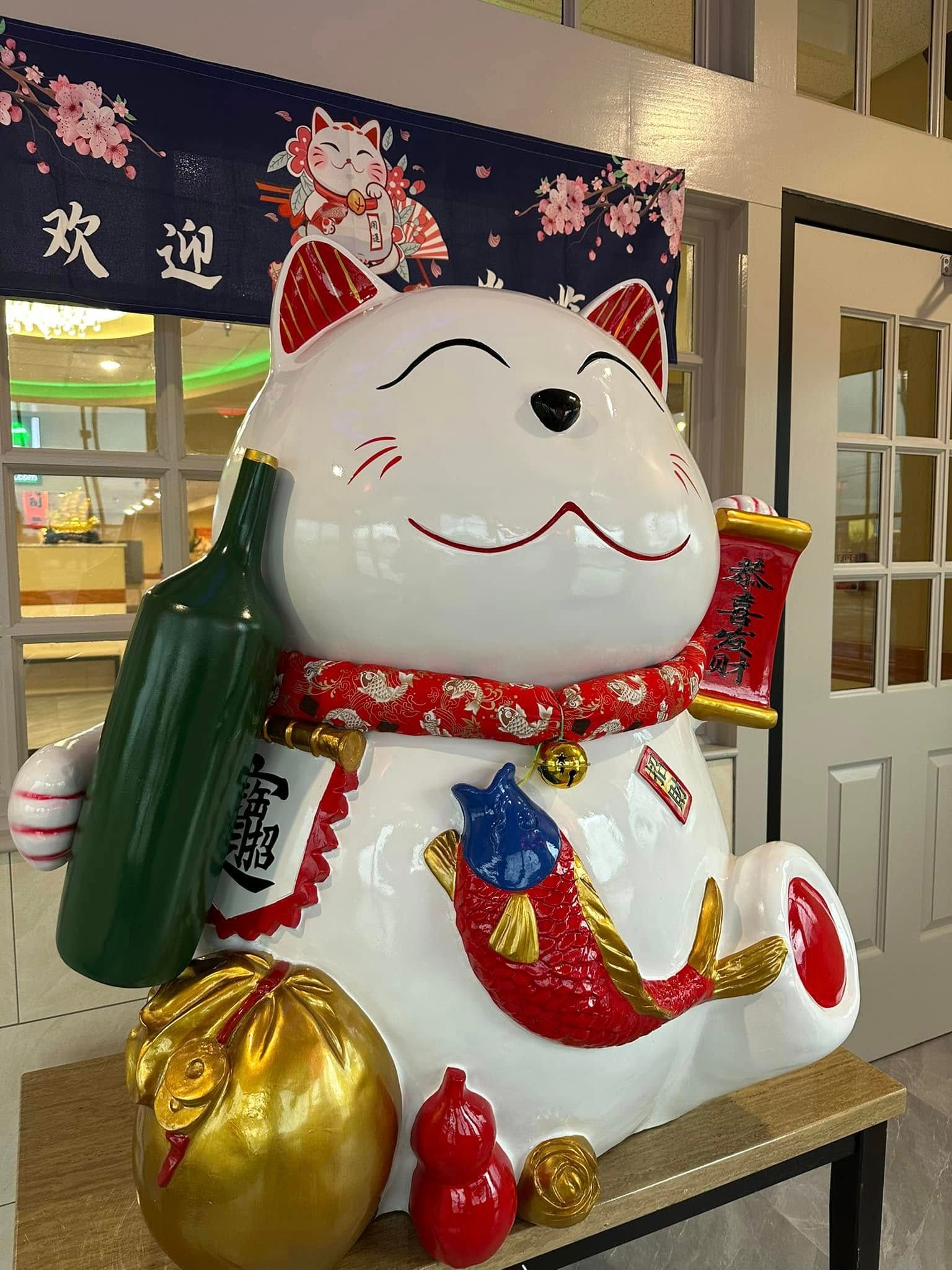 Large smiling maneki-neko statue with red scarf and gold ingots in a festive indoor lobby