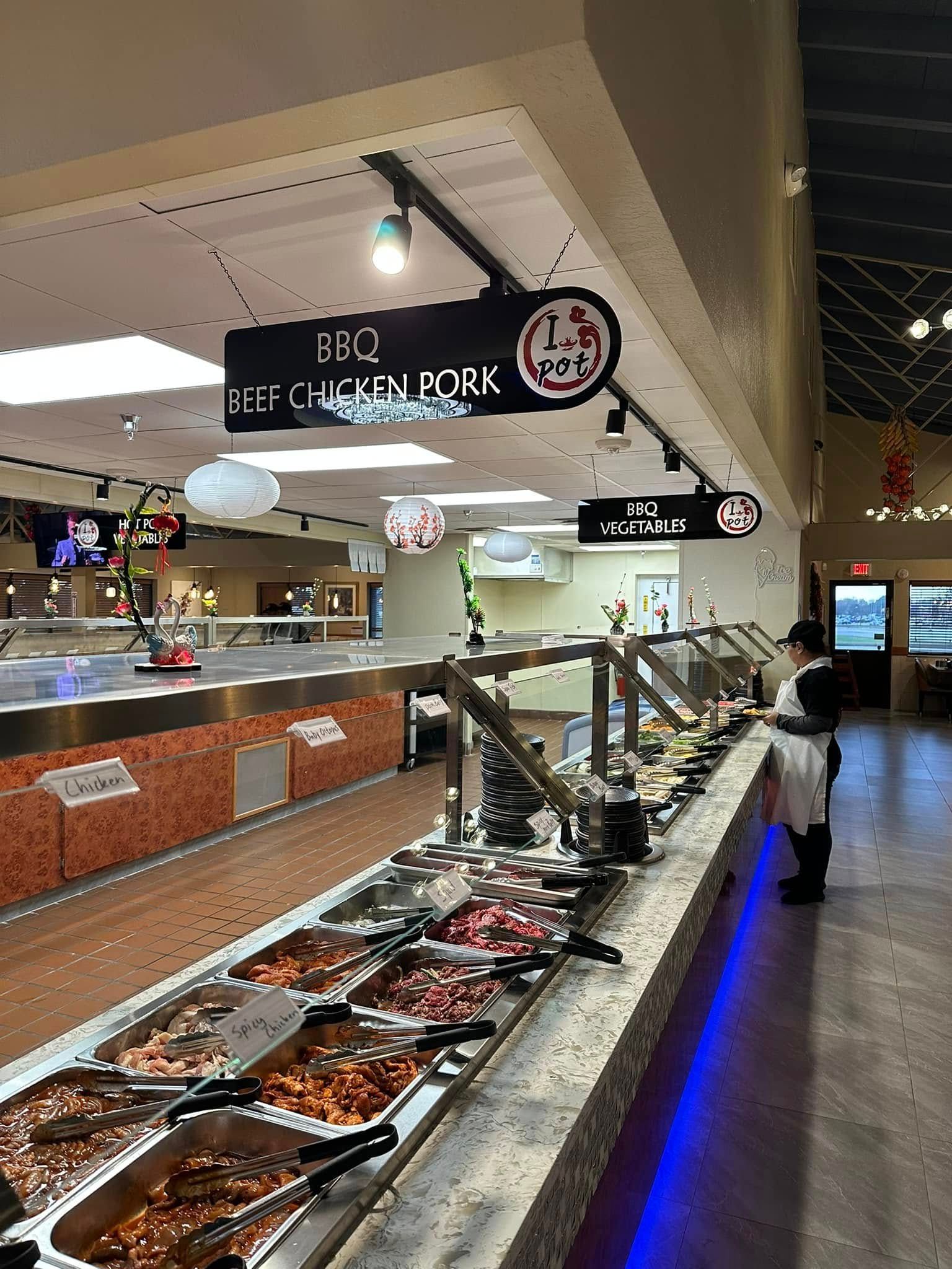 Self-serve buffet with hot food trays and hanging restaurant signs in a bright indoor dining hall