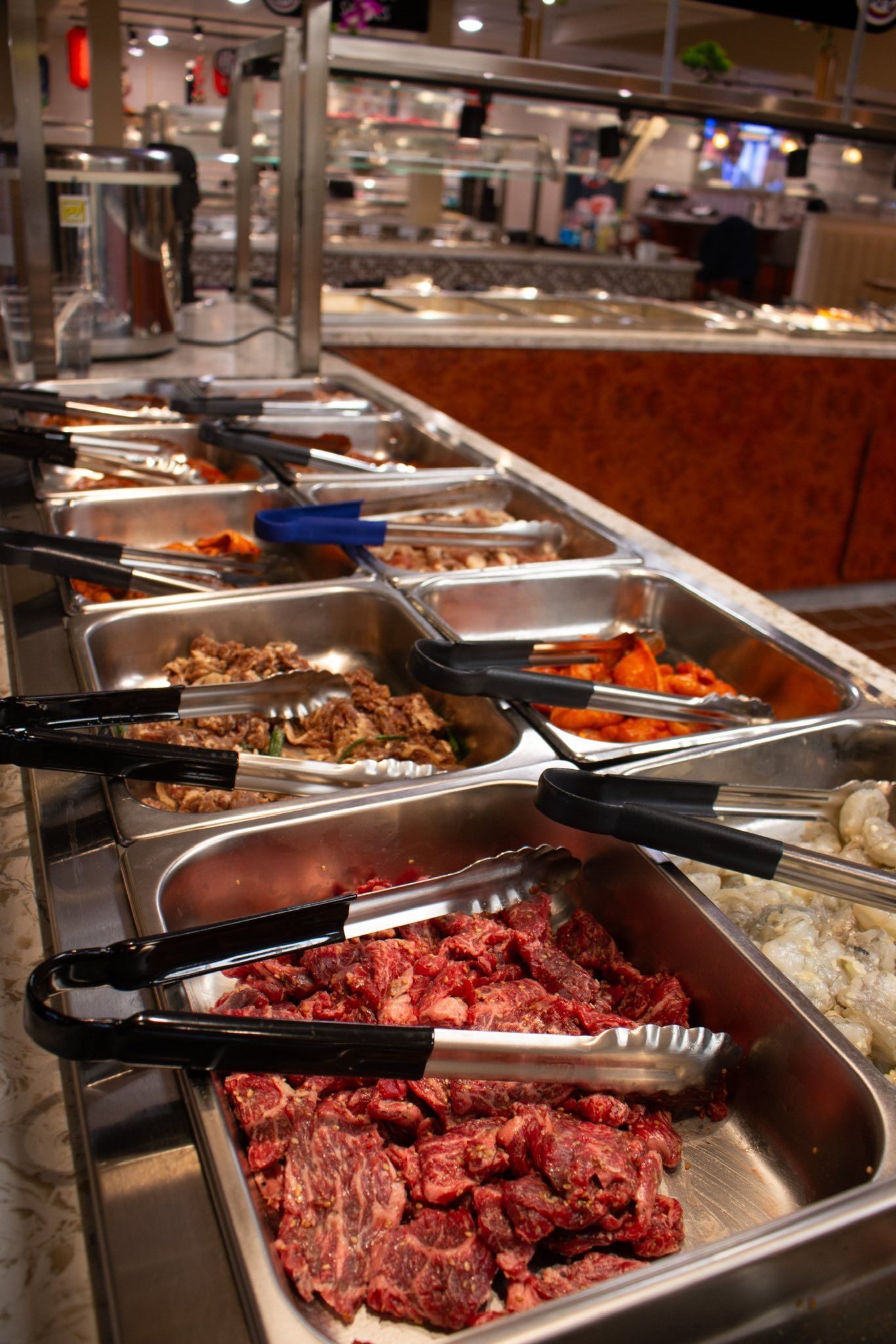 Trays of prepared meats and sides in a buffet line with serving tongs