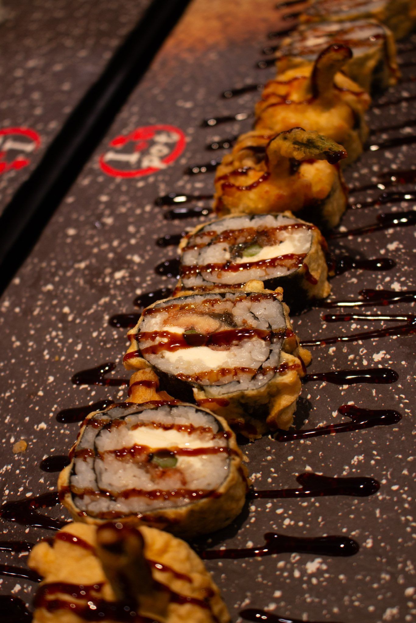 Sushi rolls drizzled with dark sauce on a textured serving tray, arranged in a diagonal line.
