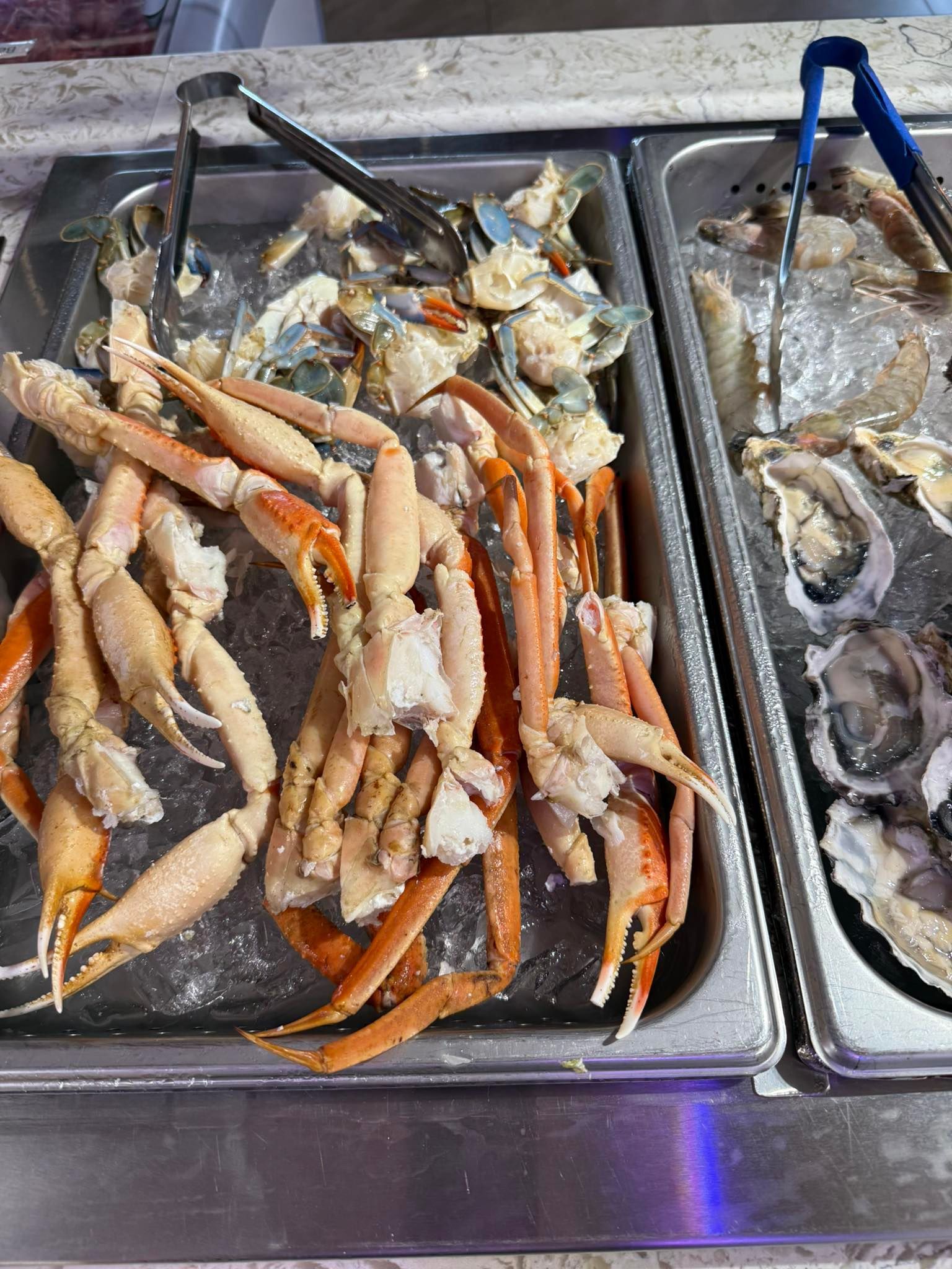 Tray of crab legs and seafood on a buffet counter, with metal tongs and a frosty display.
