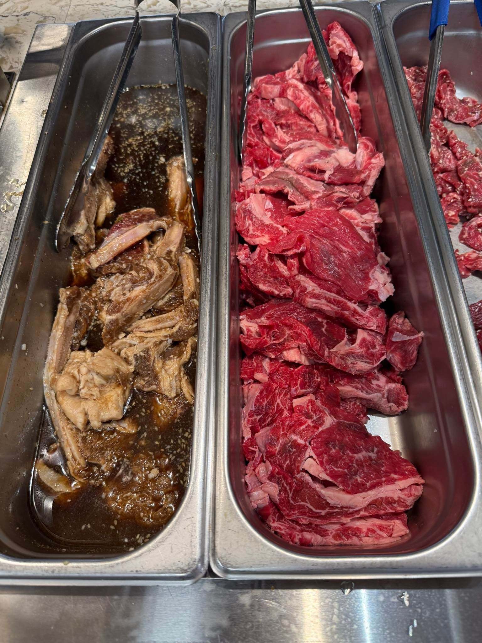 Two metal trays of marinated meat in a buffet, one brown and one red, with tongs beside them