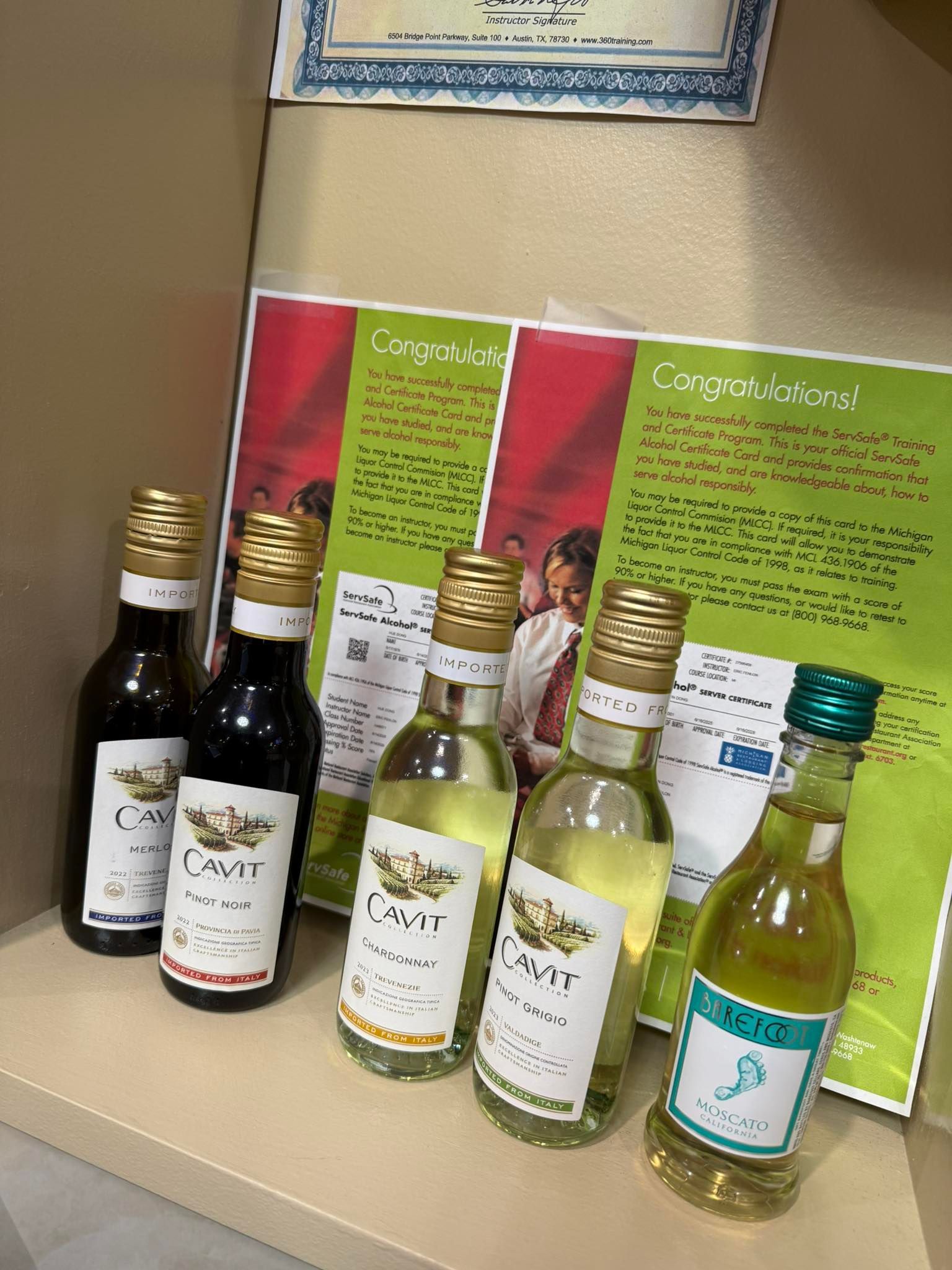 Six small bottled beverages with gold caps on a shelf, with green and red product cards behind them