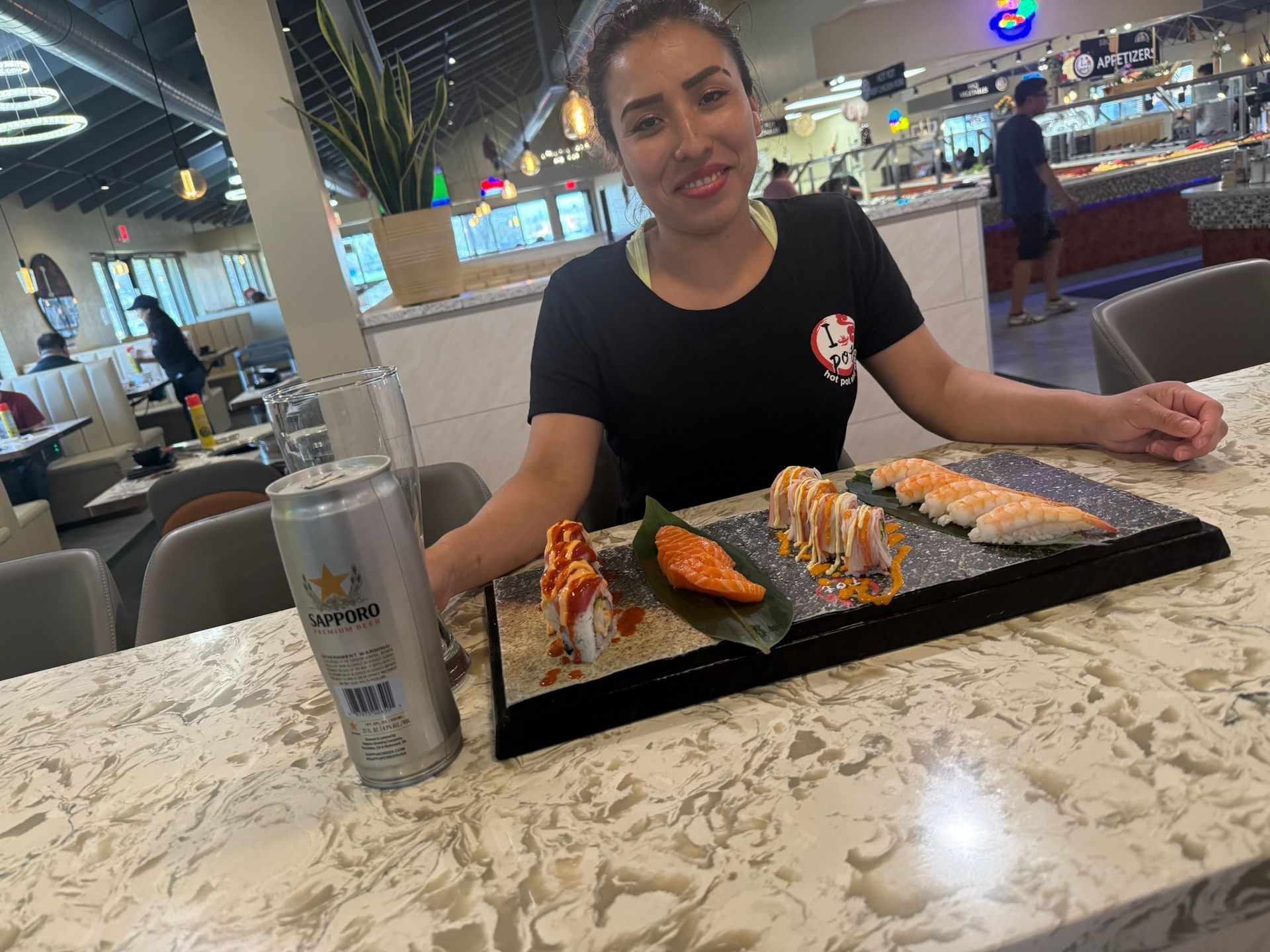 Woman seated at a sushi restaurant, smiling beside a tray of assorted sushi and a drink.