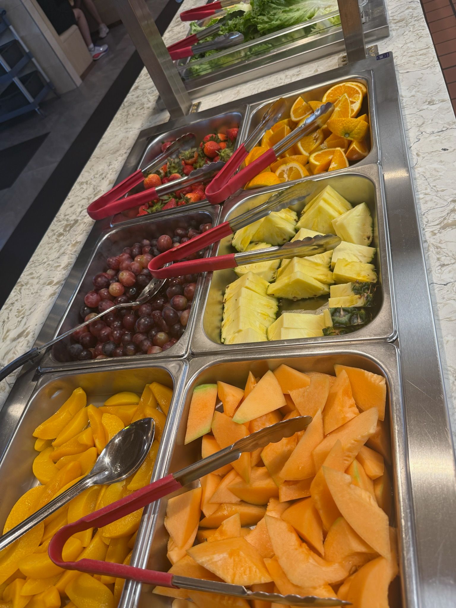 Trays of assorted cut fruit with tongs on a buffet counter, including grapes, pineapple, papaya, and melon.