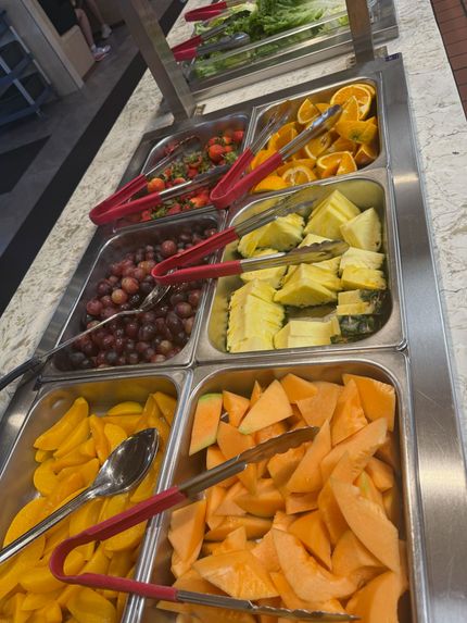 Salad bar with trays of fruit and vegetables, including grapes, pineapple, melon, and cantaloupe, with tongs.