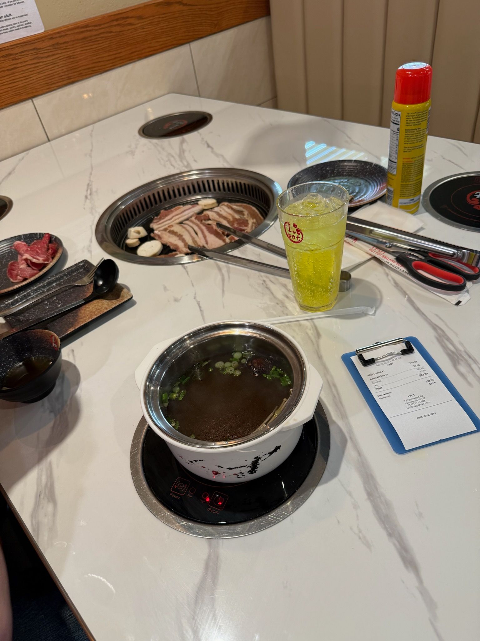 Hot pot table with bubbling broth, sliced meat, tongs, dipping sauces, and a yellow drink in a restaurant.