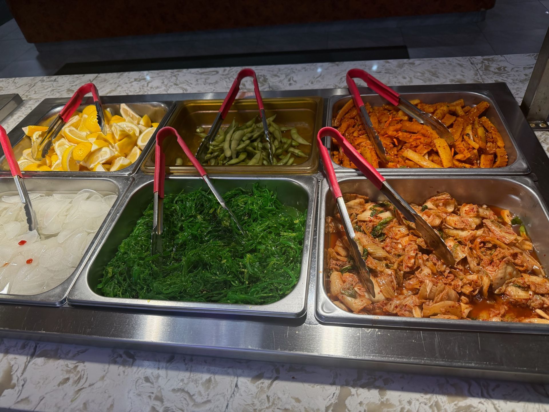 Buffet trays with assorted dishes, including rice, greens, roasted vegetables, and meat, with serving tongs.