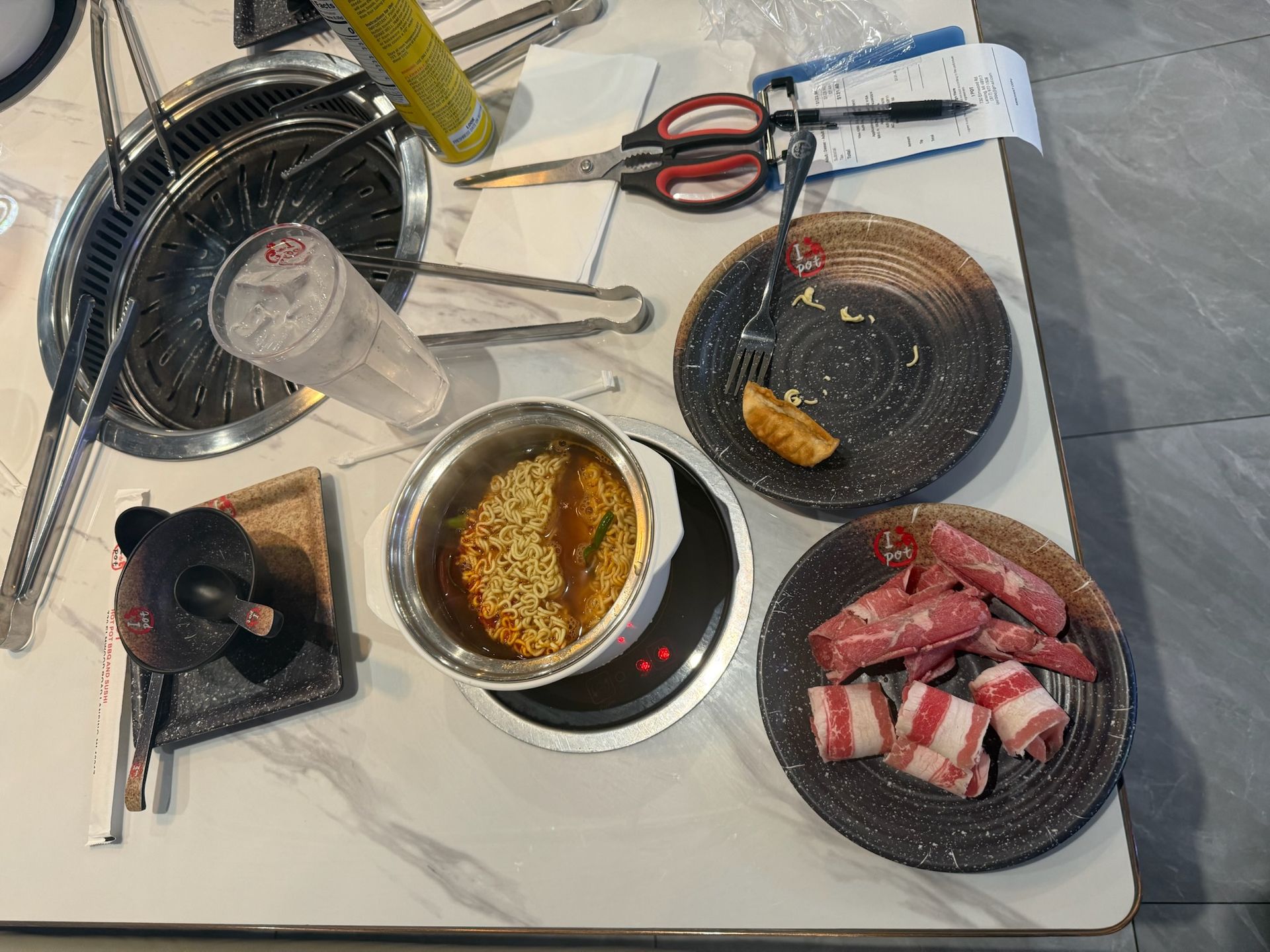 Korean barbecue table with grill, noodles, sauces, tongs, scissors, and raw meat ready to cook