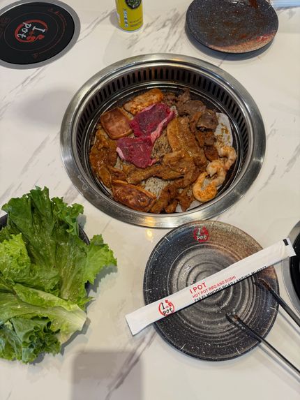 Korean barbecue grill with meat cooking, leafy greens, and dipping dishes on a table.