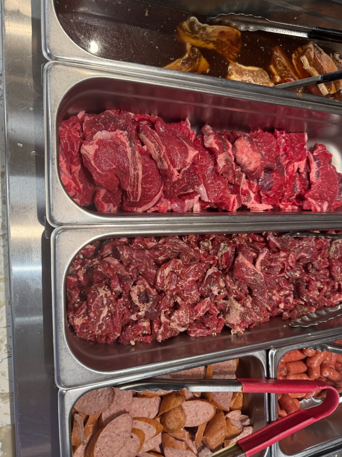 Trays of raw red meat and sausages in a stainless steel buffet display with tongs