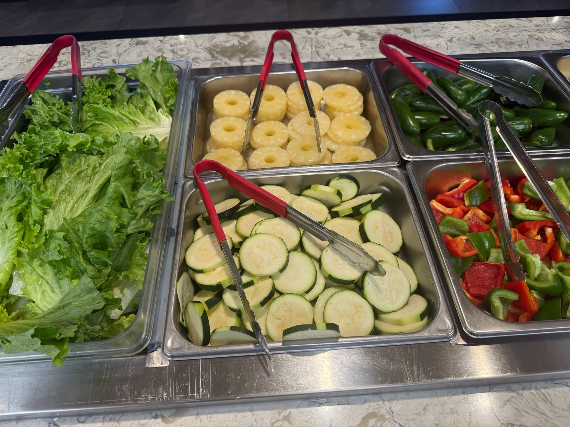 Salad bar with fresh lettuce, zucchini, pineapple rings, and bell peppers in metal trays with tongs