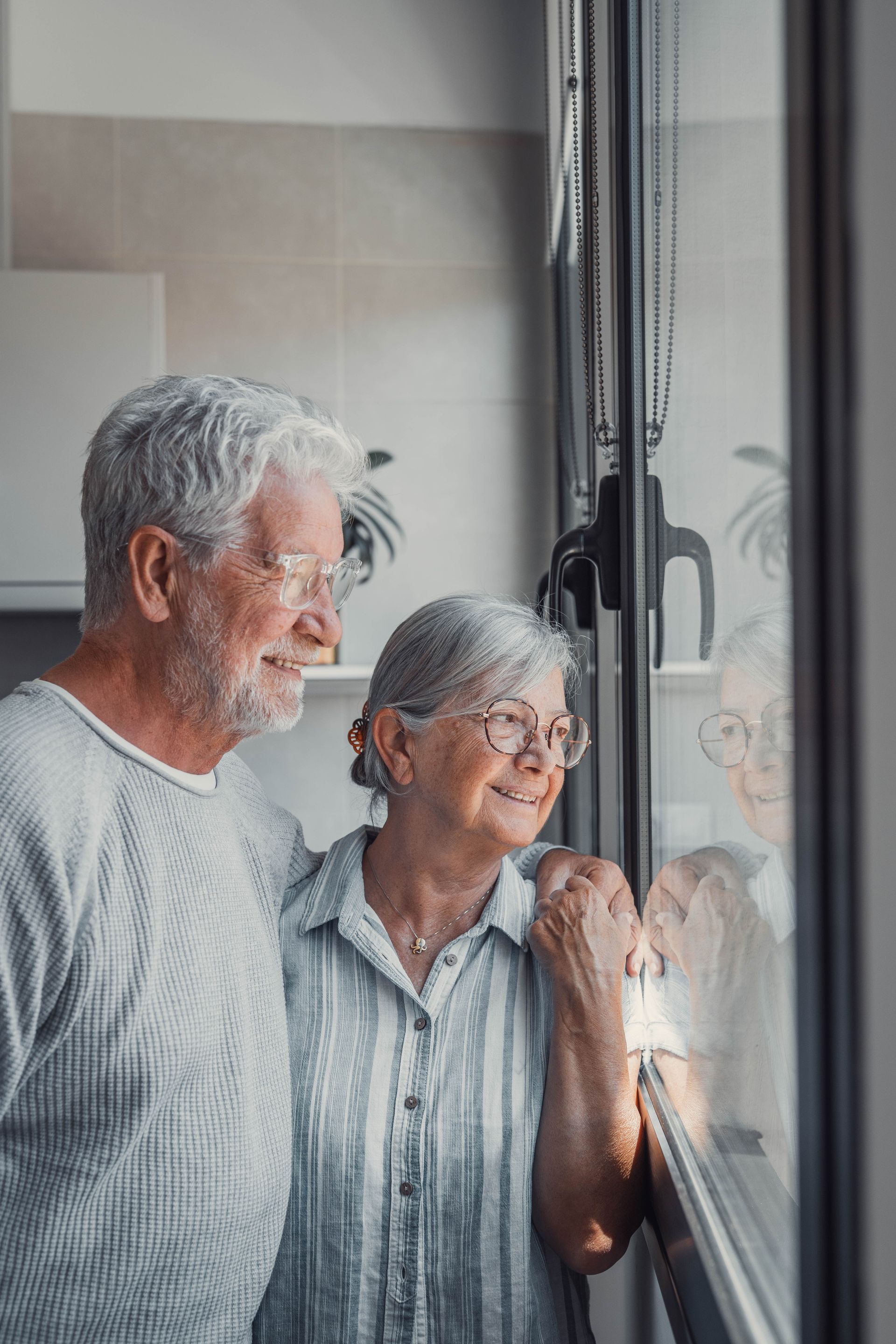 Two older adults stand by a window, looking outside together in a bright room.