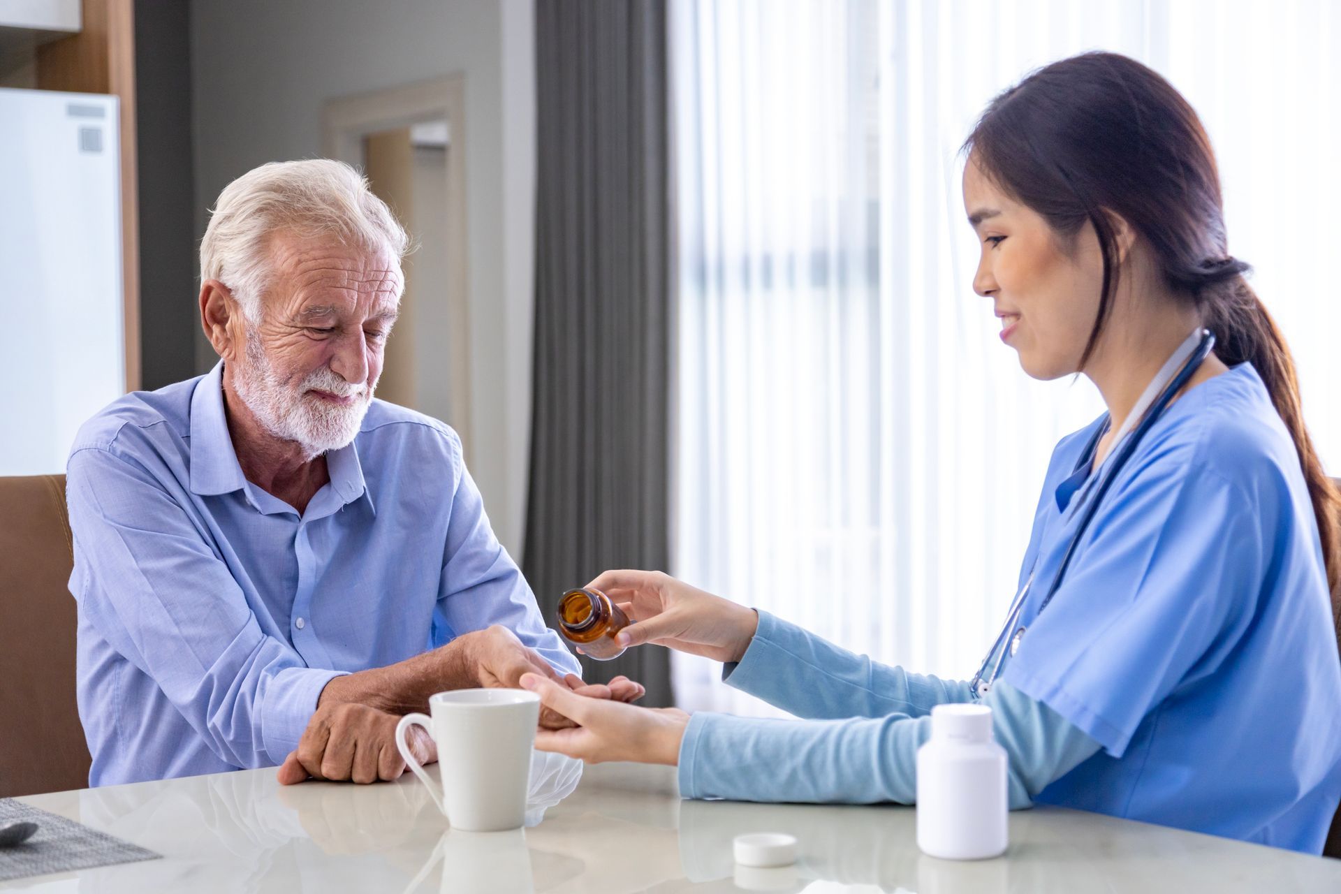 Caregiver offering medicine to an elderly man at a table in a bright room