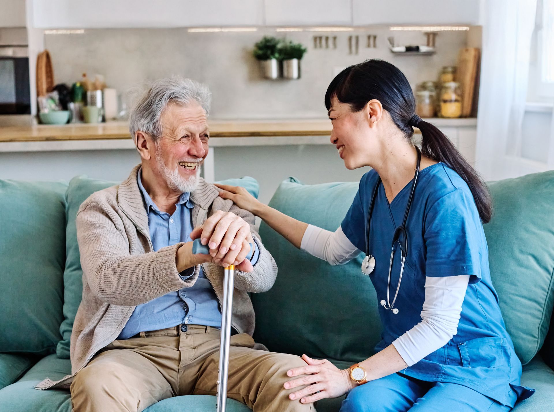 Caregiver in blue scrubs smiling and holding an older man's hand on a couch in a bright living room