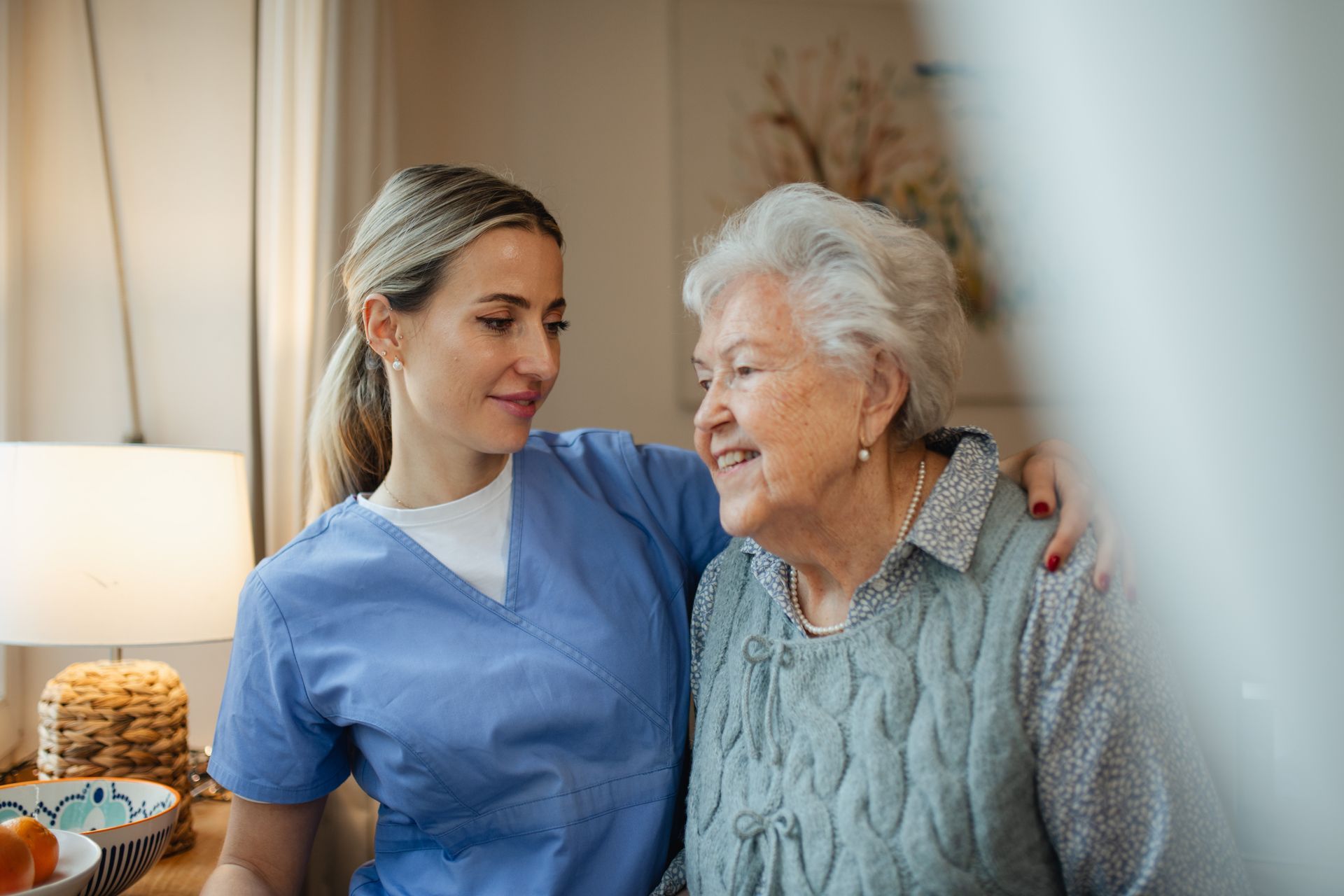 Two healthcare workers smiling in a bright room, one comforting the other with an arm around her shoulder