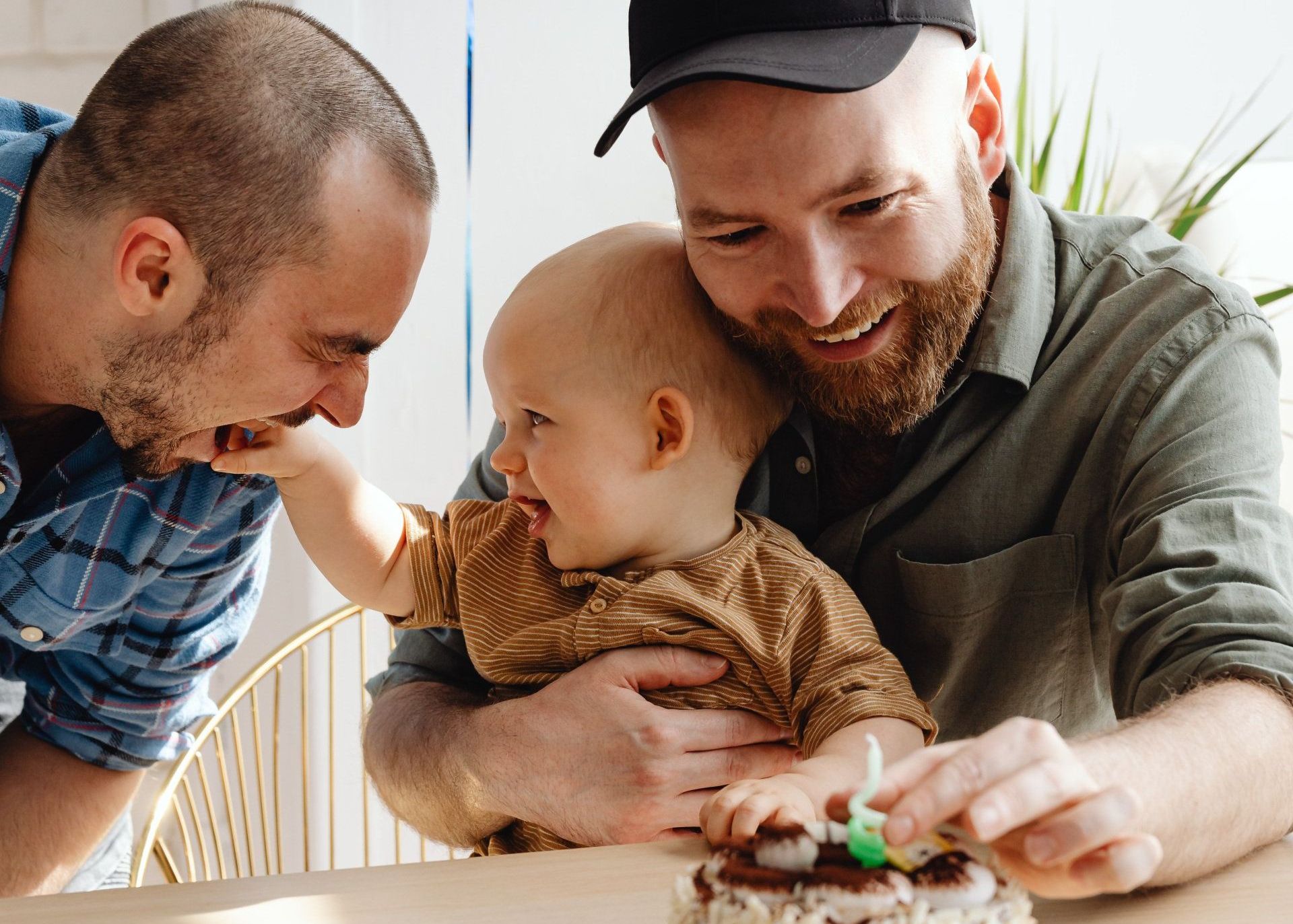 Two men are sitting at a table with a baby and a cake.