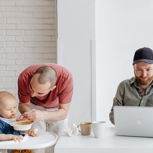 A man is feeding a baby while another man is using a laptop.