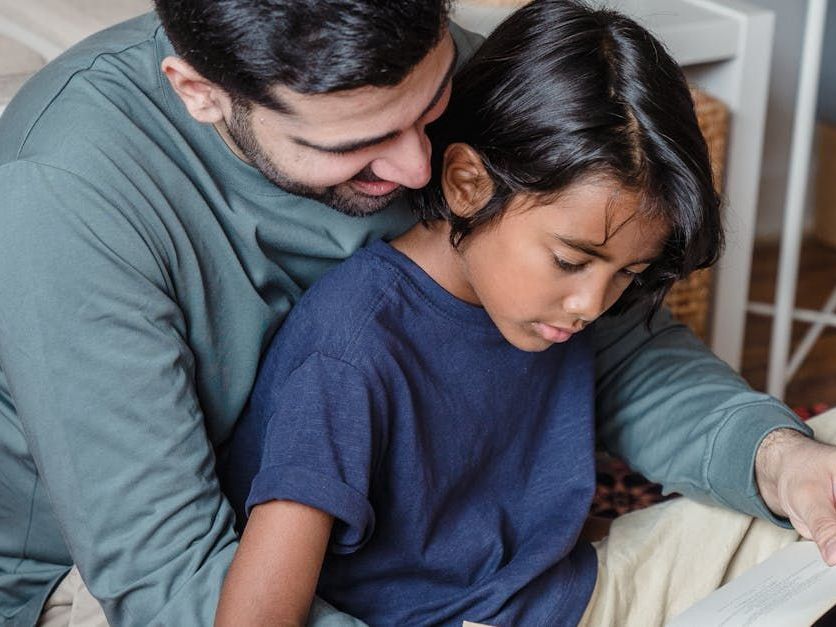A man is sitting on the floor reading a book to a young boy.