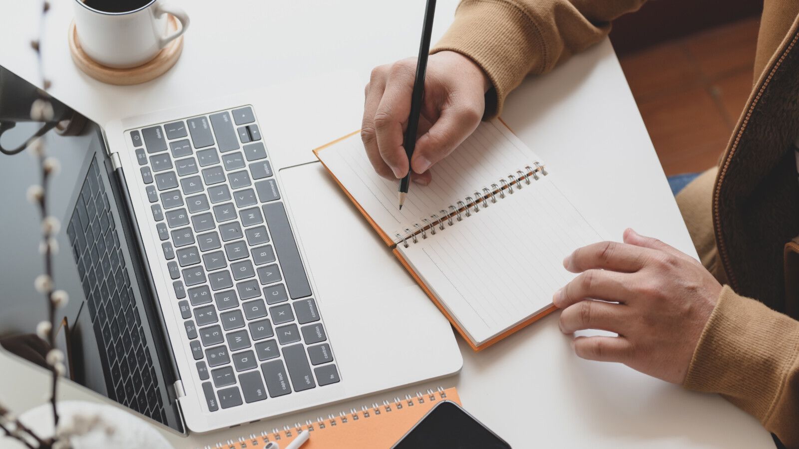 A person is writing in a notebook next to a laptop computer.