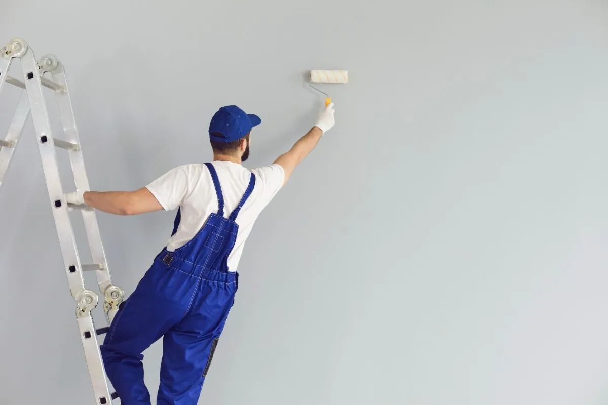 Un operaio in tuta blu dipinge un muro vicino a una scala con un rullo.
