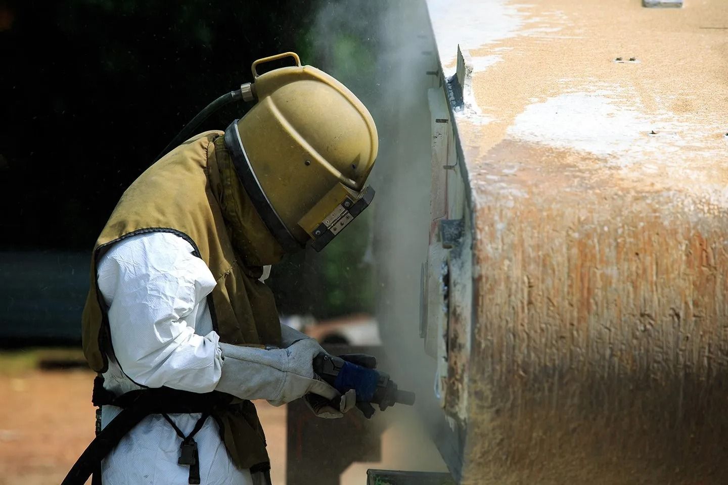 Operaio con indumenti protettivi esegue la sabbiatura di un grande serbatoio metallico all'aperto.