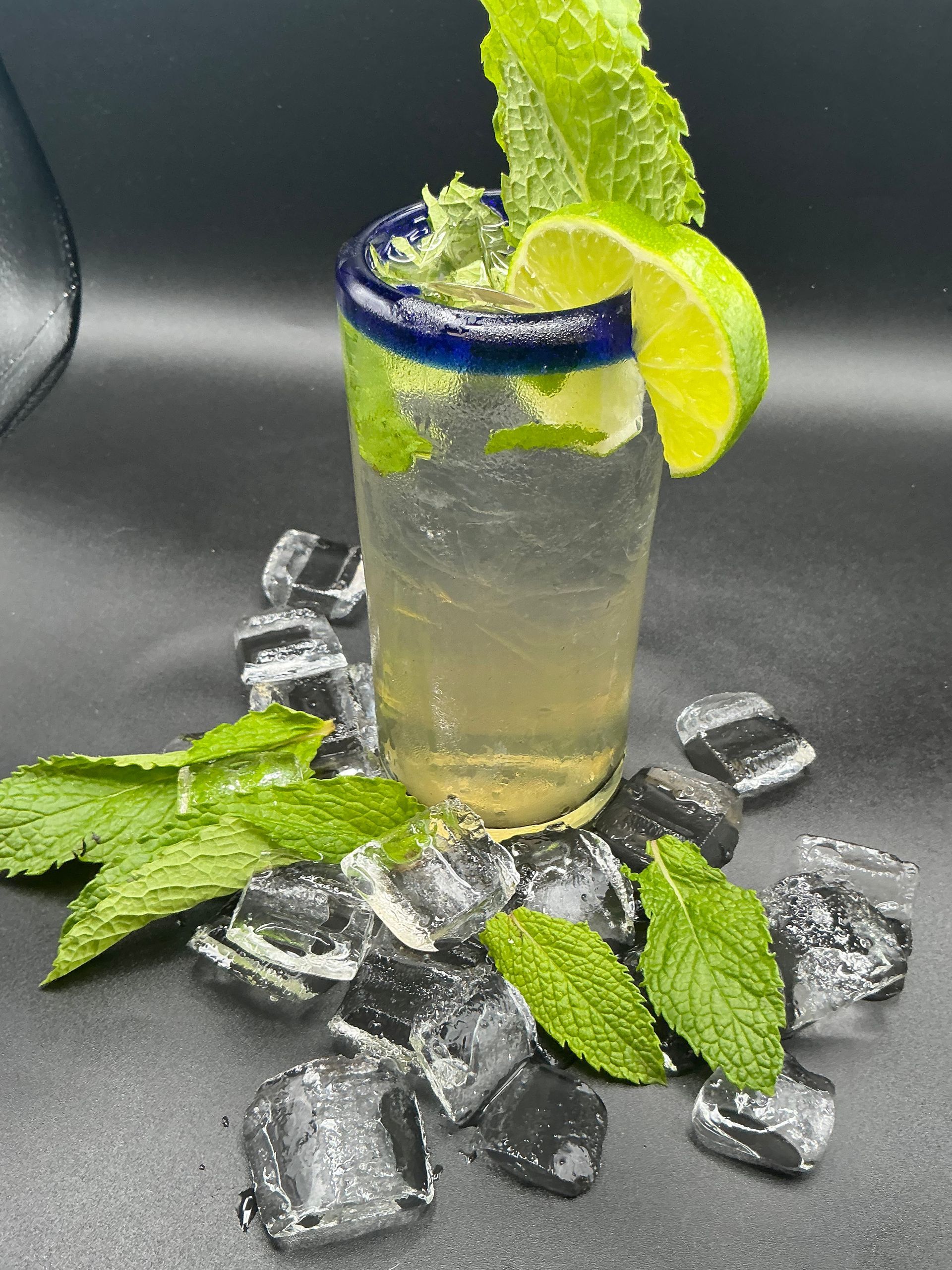 Mojito cocktail in a blue-rimmed glass, garnished with lime and mint, surrounded by ice and mint leaves.