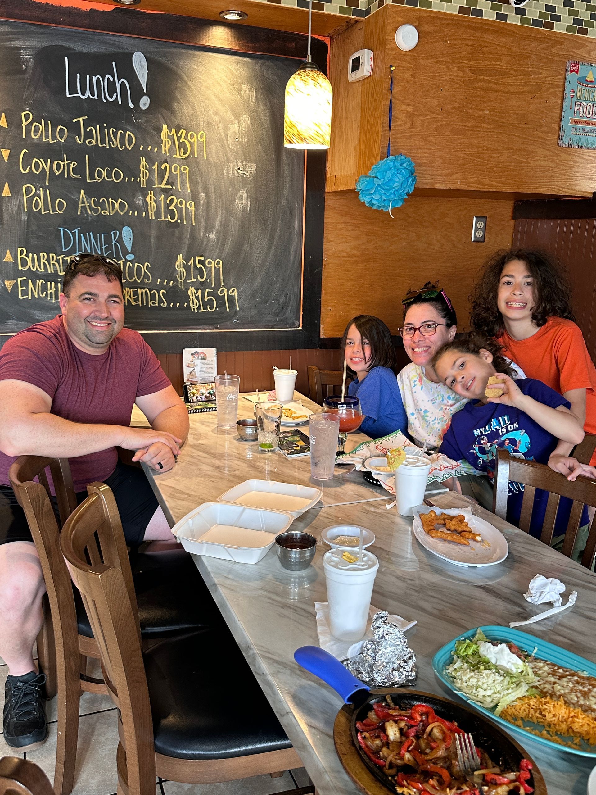 Family at a restaurant table with food. Man at left, women and children seated at right; chalkboard menu visible.