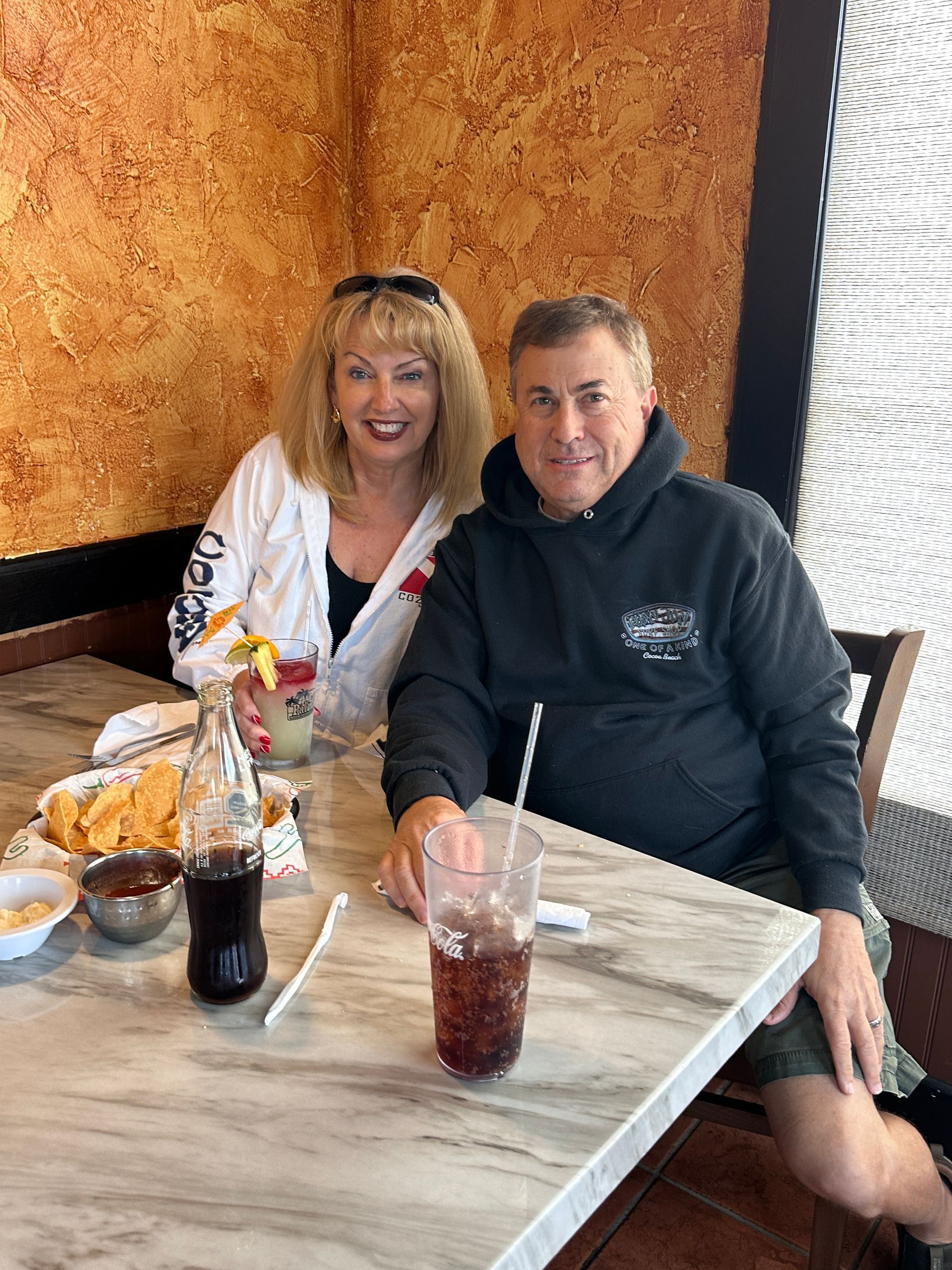 Two people at a restaurant table with drinks, chips, and salsa. Woman smiles; man holds a drink.