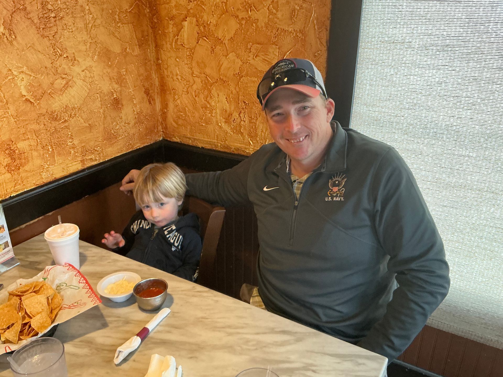 Man and child sitting at a restaurant table with chips, salsa, and drinks. The man has an arm around the child.