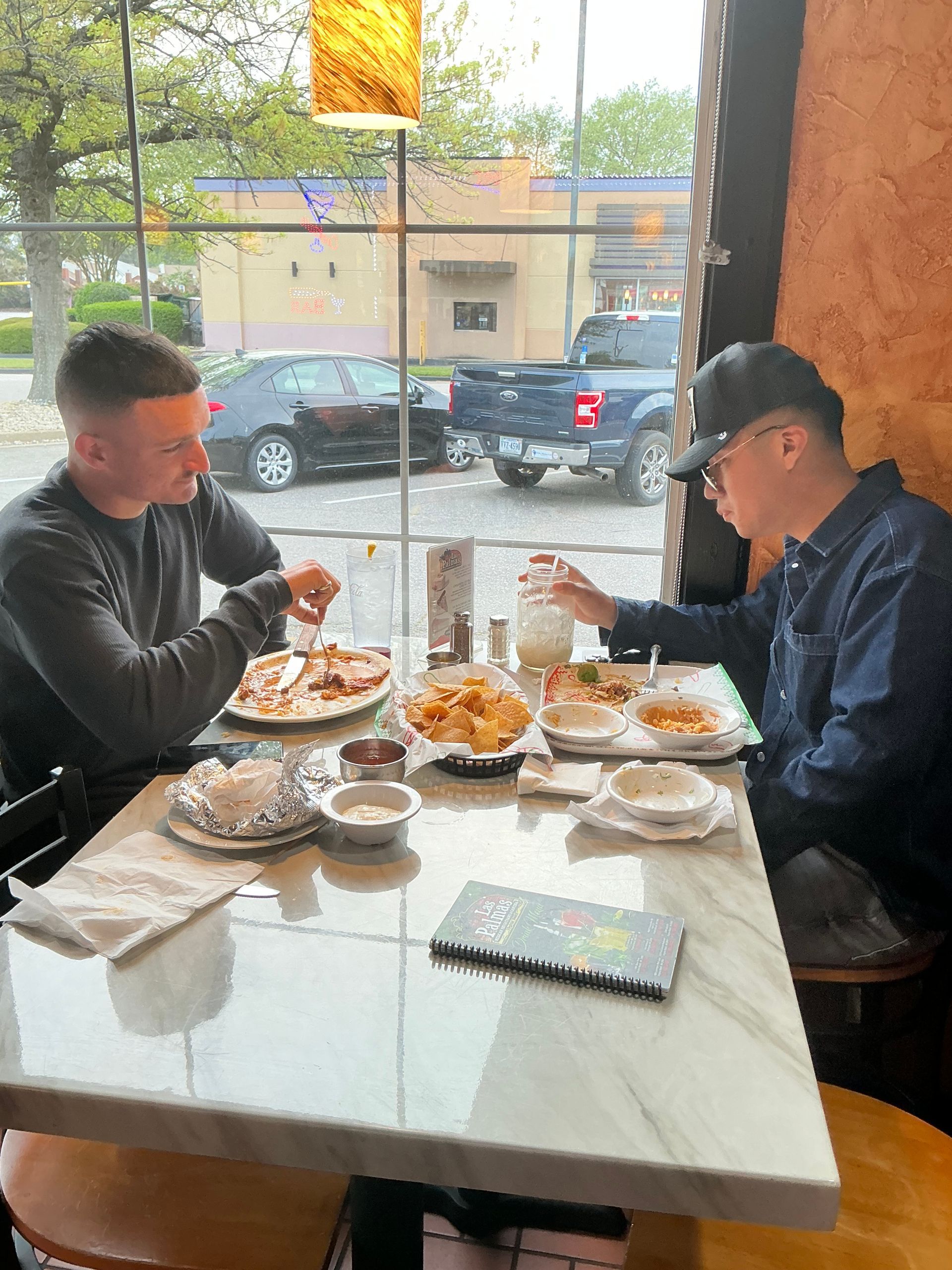 Two people eating at a restaurant table. Pizza, fries, and drinks are visible. A window is in the background.