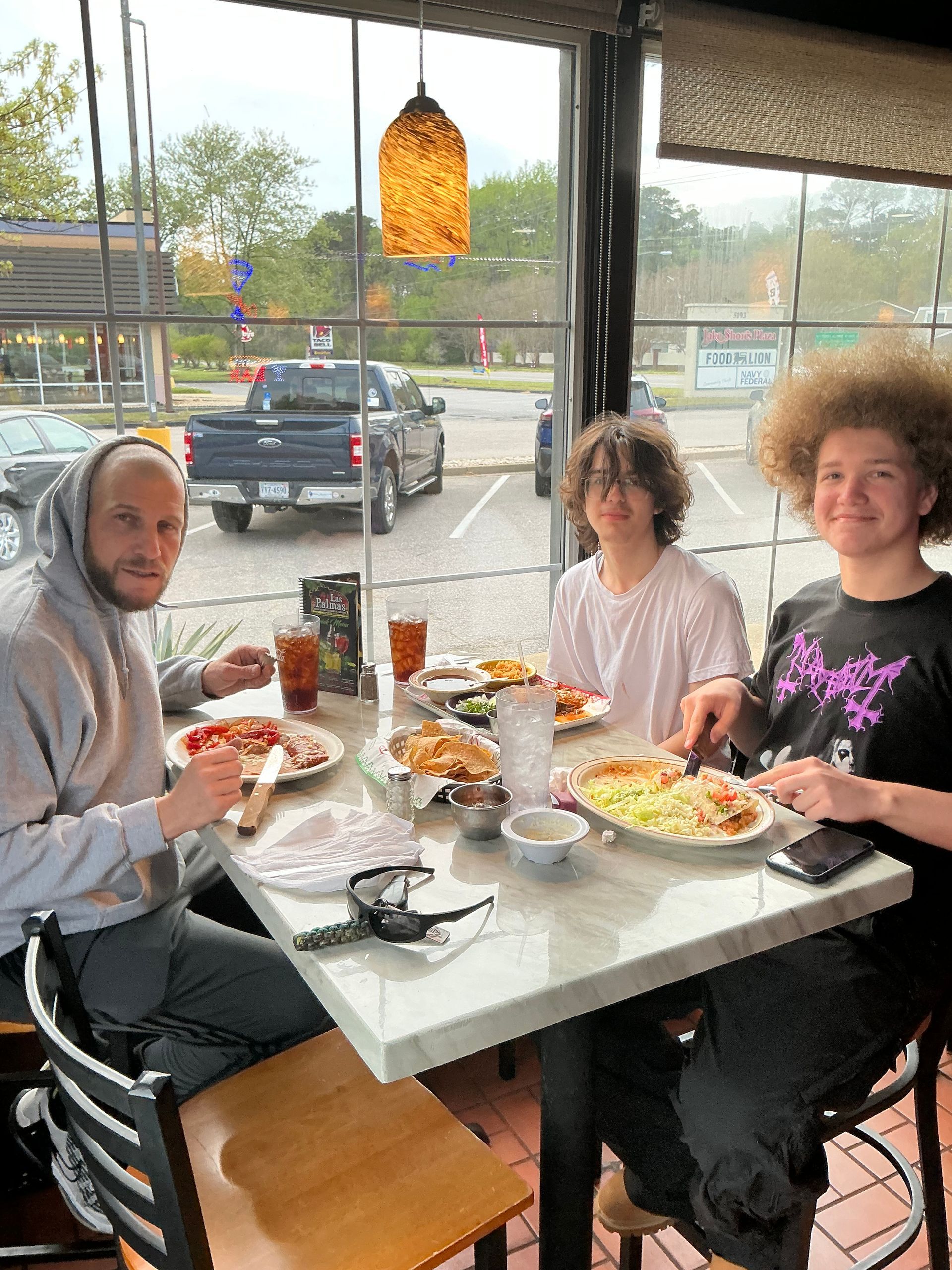 Three people at a restaurant table, eating. Bald person, two people with curly hair. Food and drinks are on the table.
