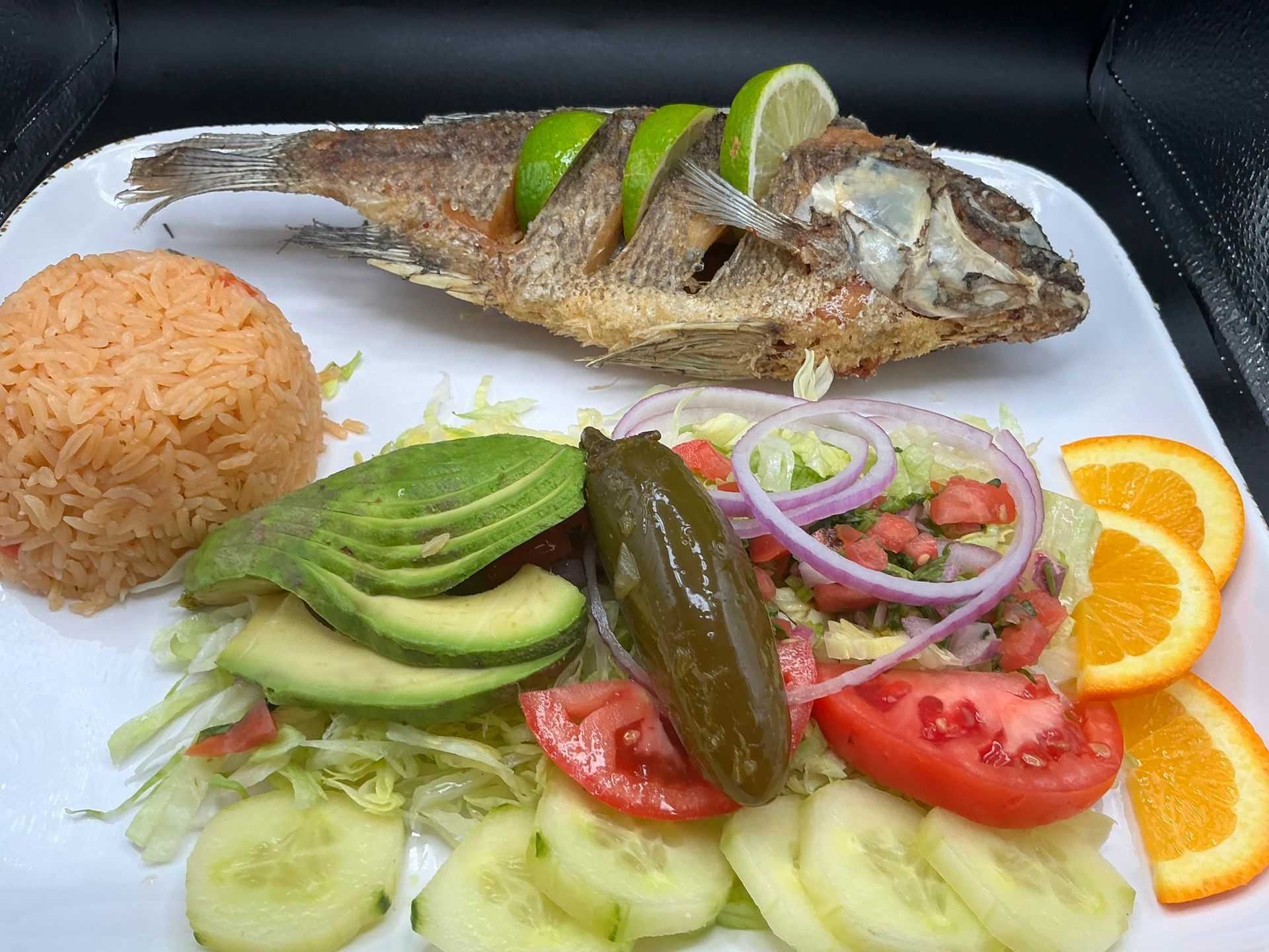 Fried fish with rice, avocado, and vegetables on a white plate, garnished with lime and orange slices.