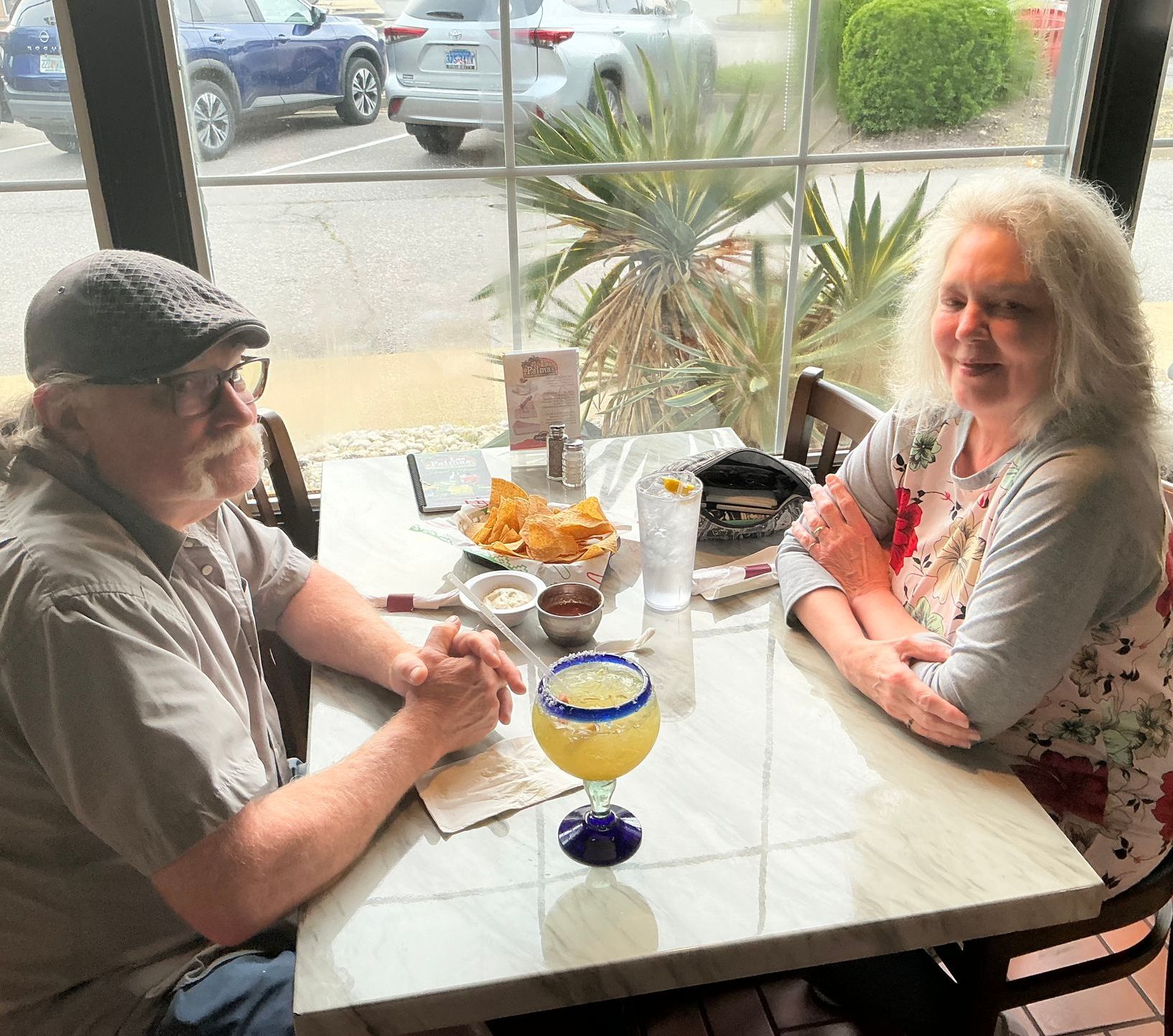 A couple sits at a restaurant table with chips, salsa, and a margarita by a window.