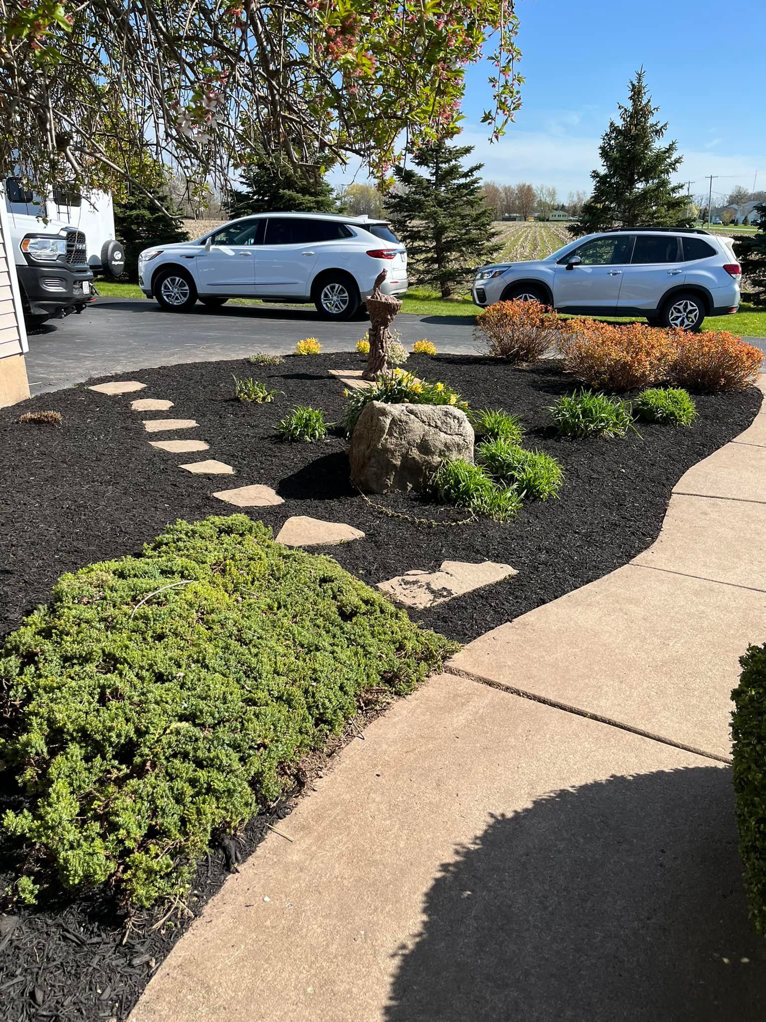 Curved landscaped garden bed with mulch, stepping stones, shrubs, and parked cars beside a sidewalk